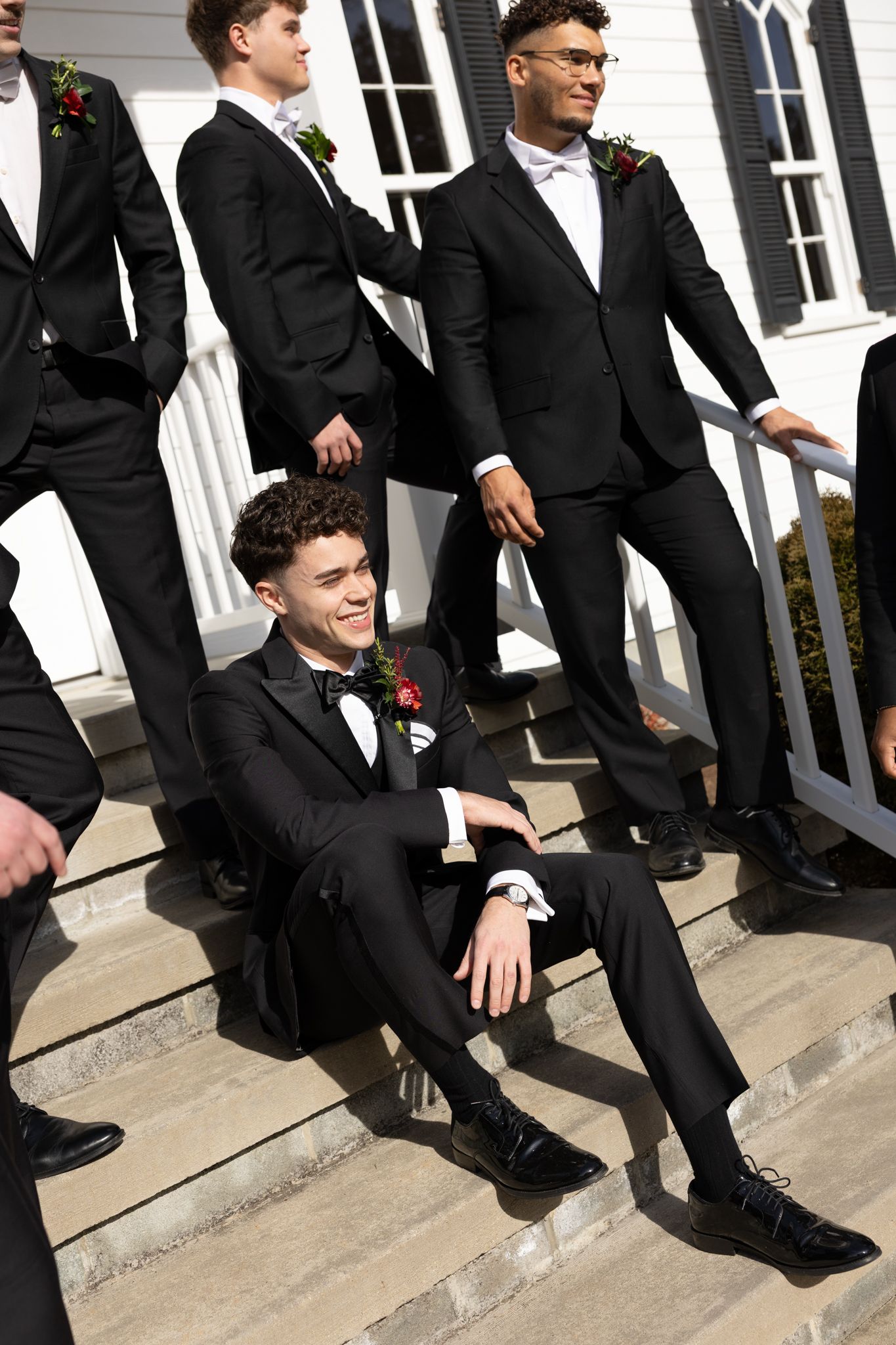 Young groomsmen in black tuxedos with red boutonnieres pose on white church steps in sunny outdoor wedding photo.