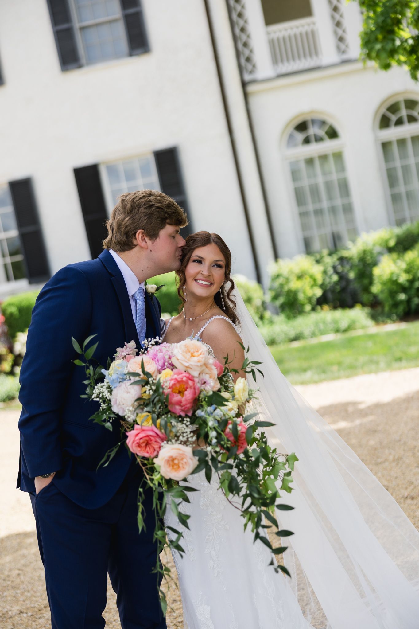 A groom in a navy suit kisses his bride on the cheek as she smiles holding a lush colorful bridal bouquet.