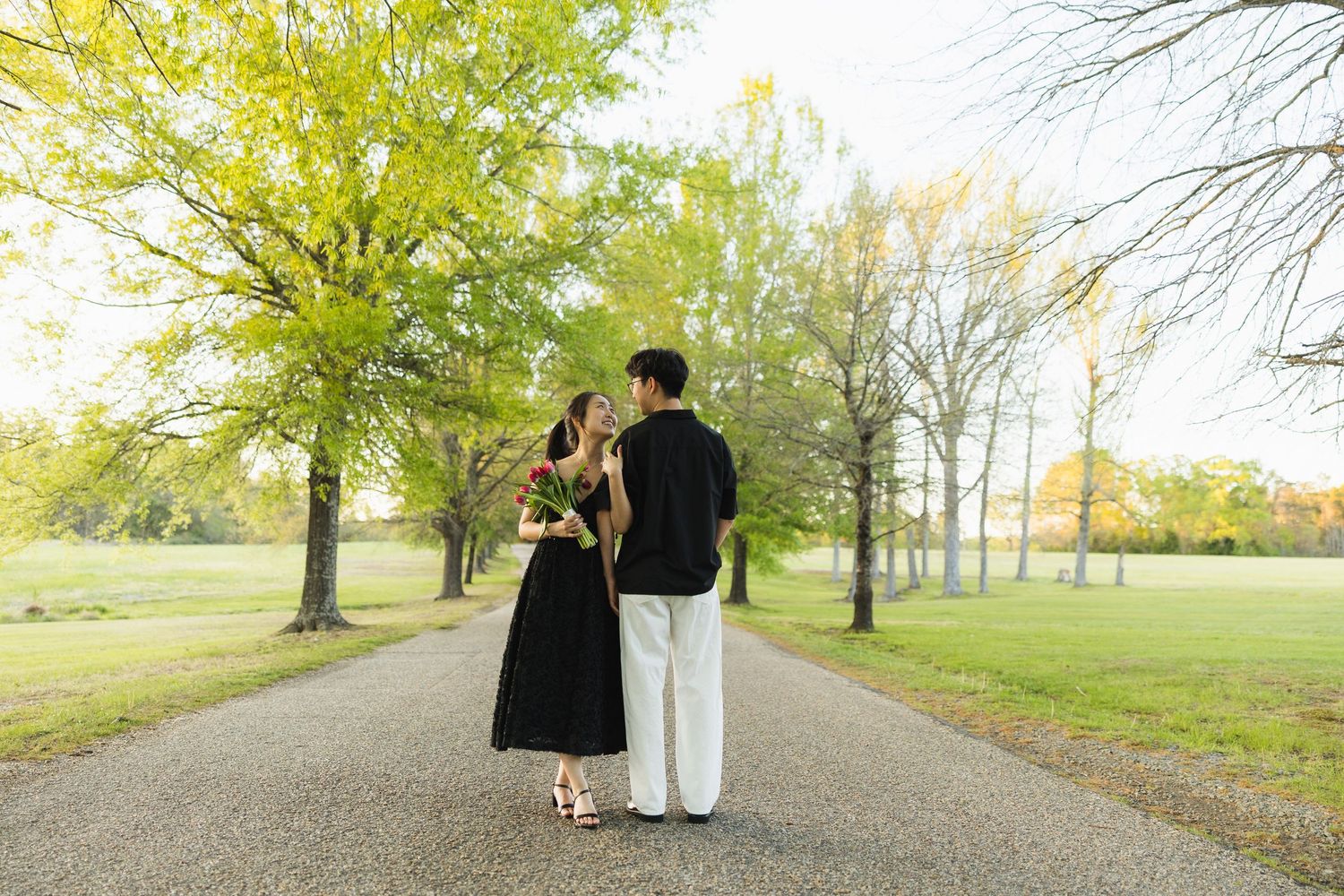 A couple in black outfits walks hand in hand down a tree-lined path, woman holding red roses.