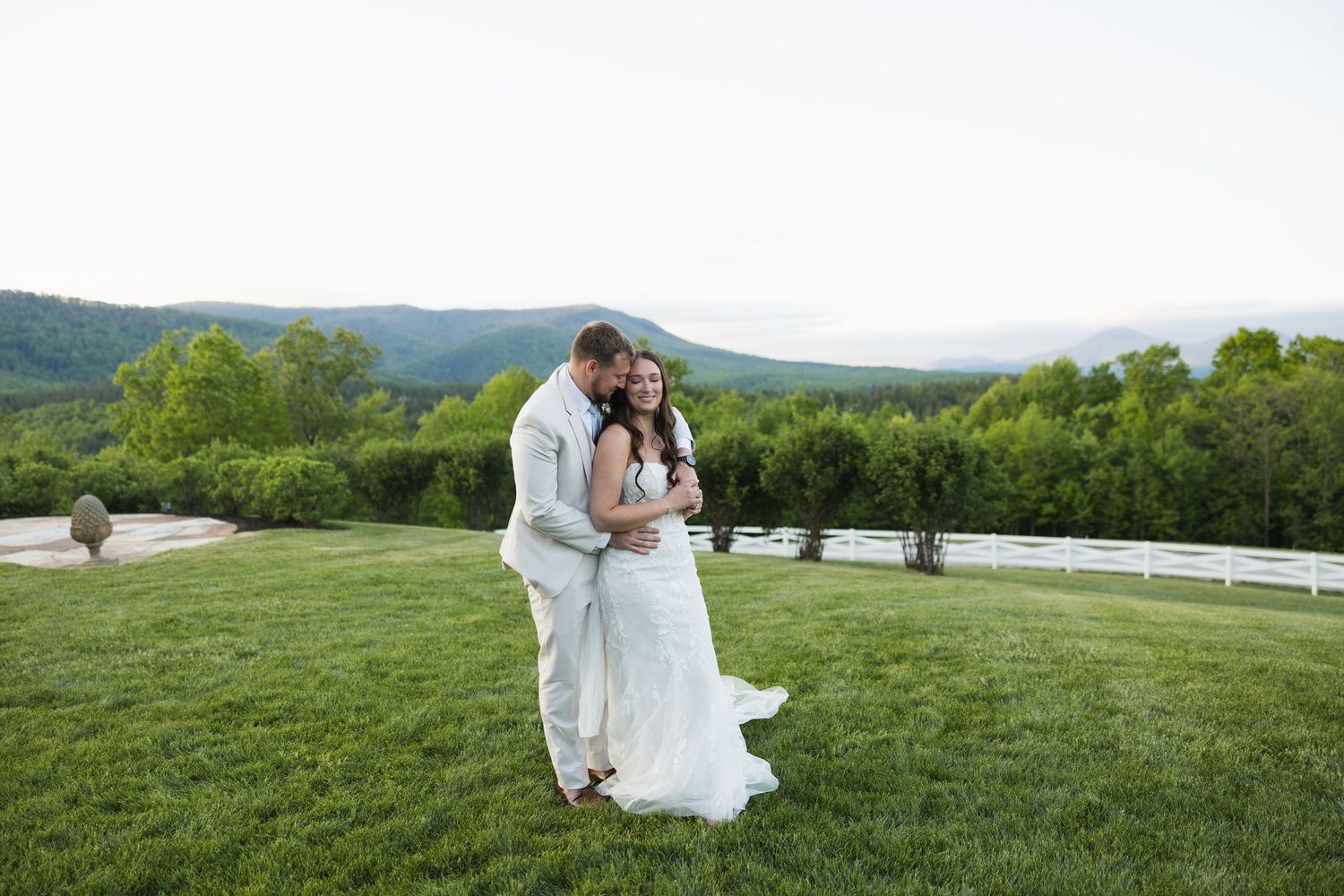 Happy bride and groom embrace on green lawn with scenic mountain views at sunset.