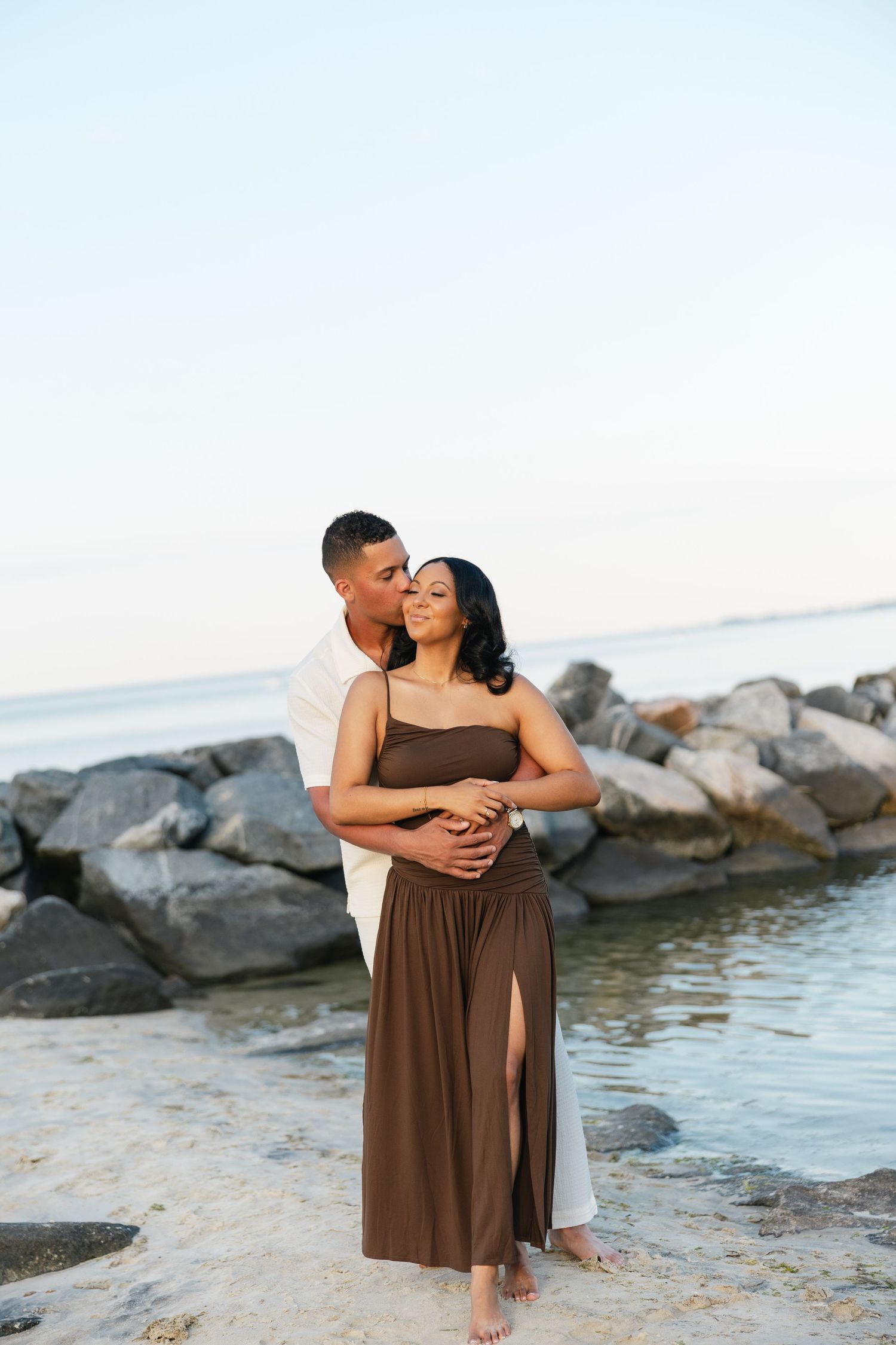 Couple embracing on a rocky beach, man in white shirt kissing woman in brown maxi dress at sunset.