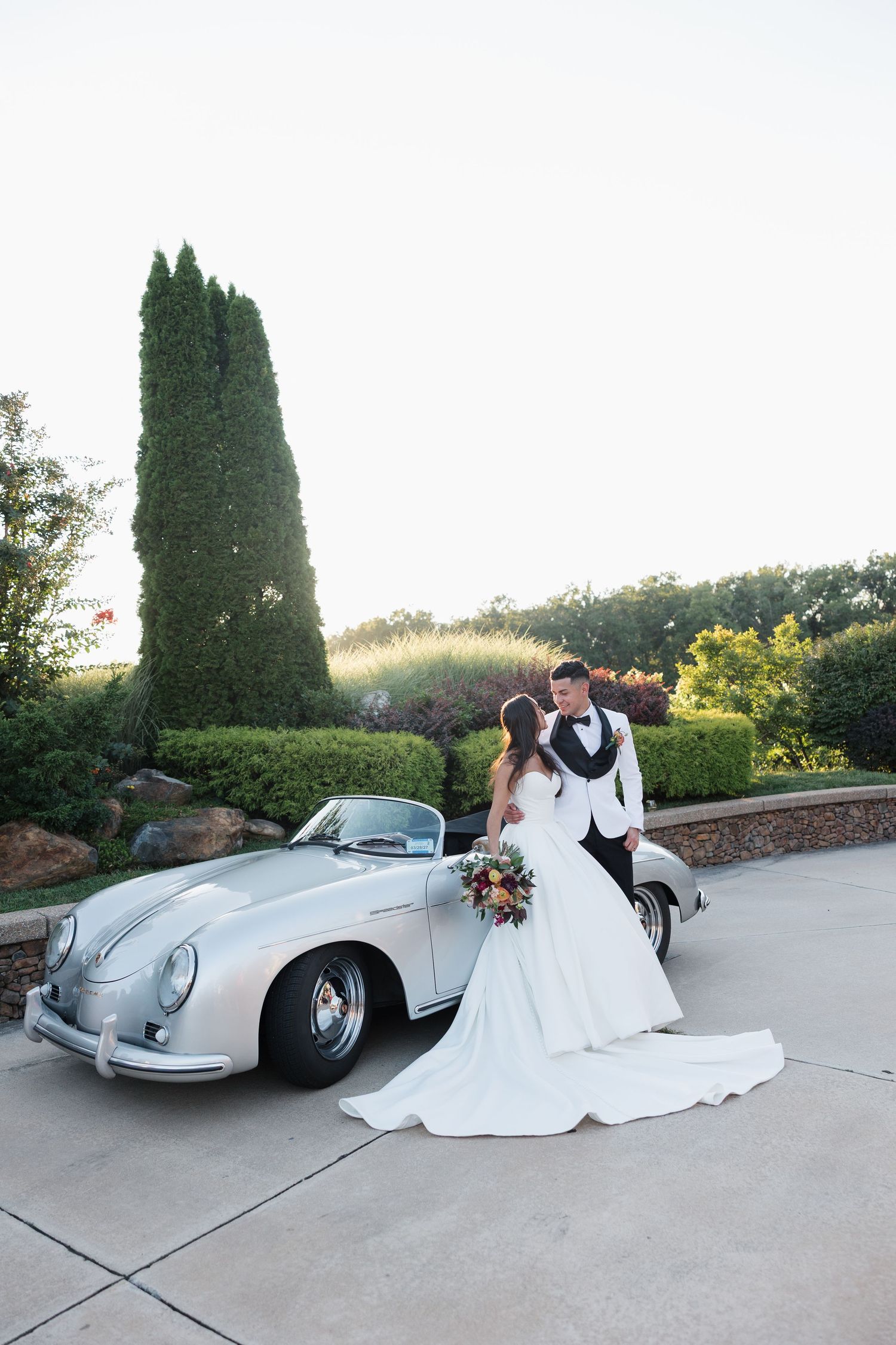 Bride and groom pose romantically beside a classic silver Porsche convertible in a lush garden setting at golden hour.