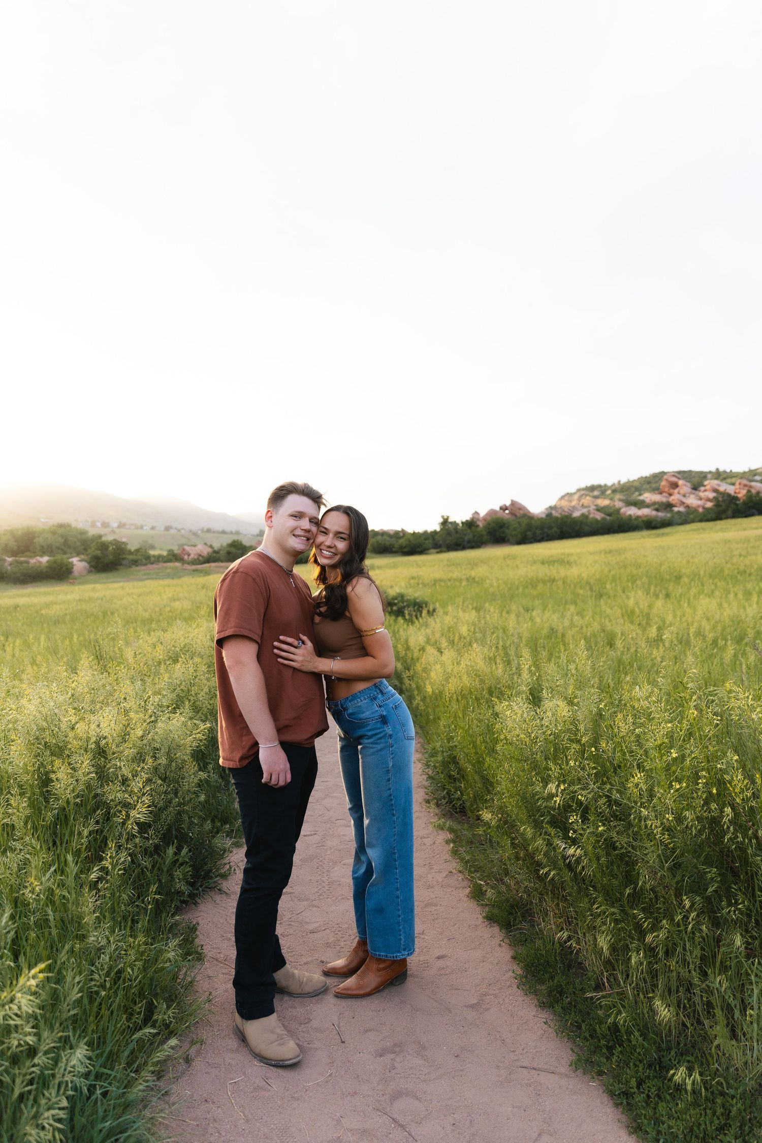 Smiling couple embracing on a dirt path through a lush green field at golden hour.