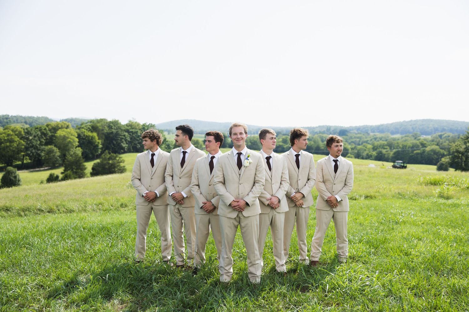 Groomsmen in matching beige suits and navy ties stand in a lush green field with rolling hills backdrop.
