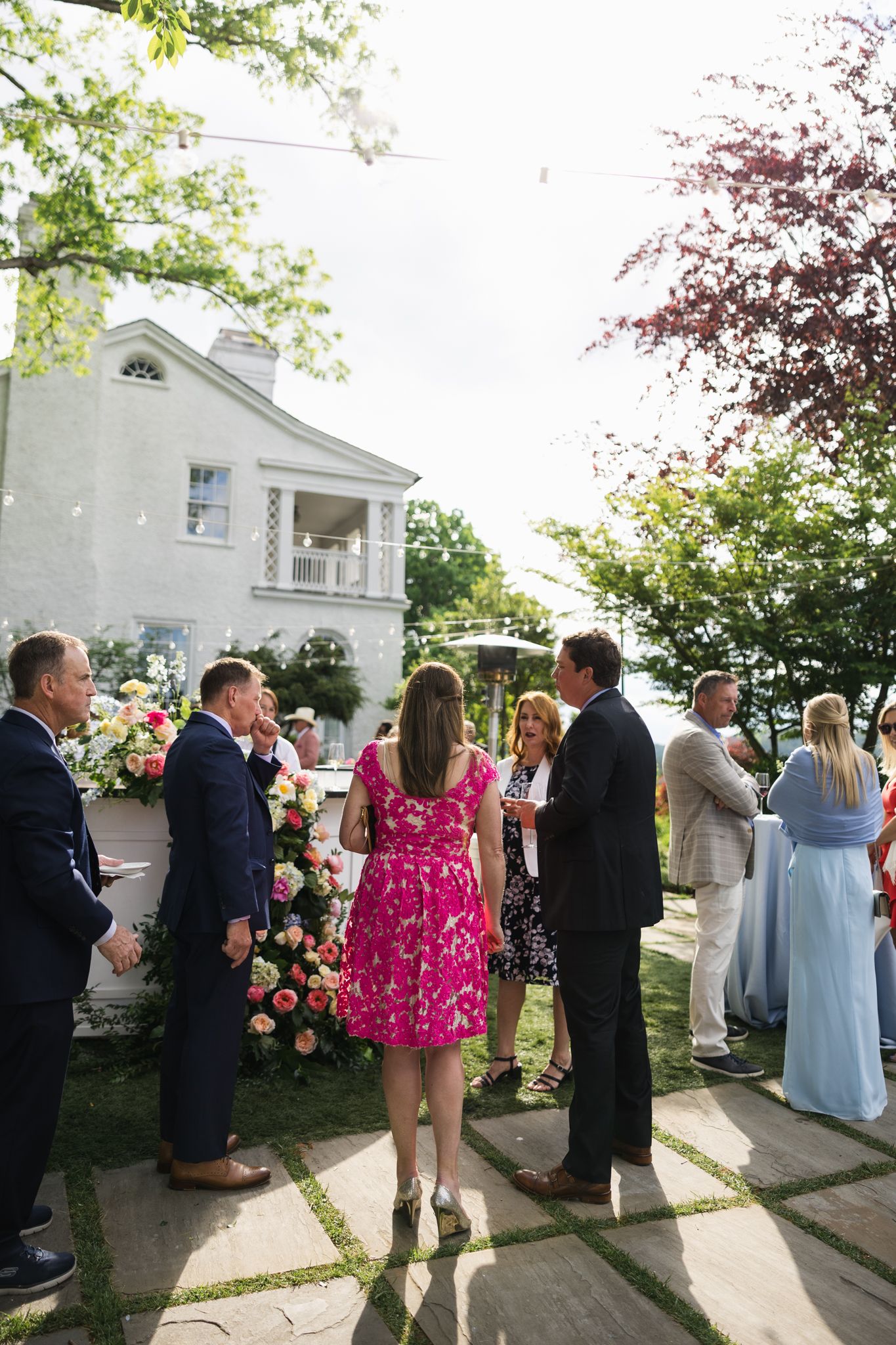 Guests mingle outdoors at an elegant garden wedding reception near a white colonial home with colorful floral arrangements.