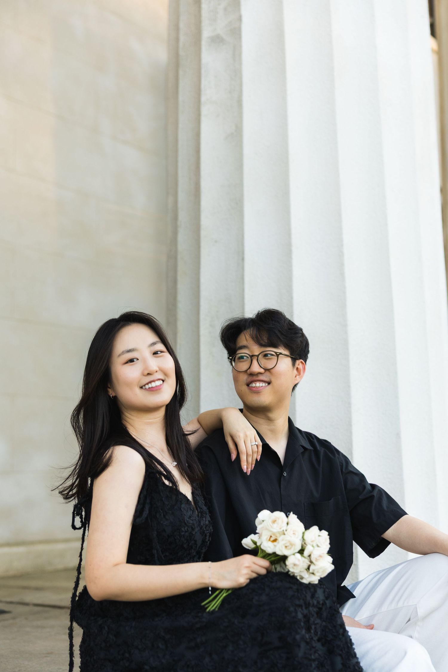 A happy couple poses together by white columns, woman holding white flowers and smiling brightly.
