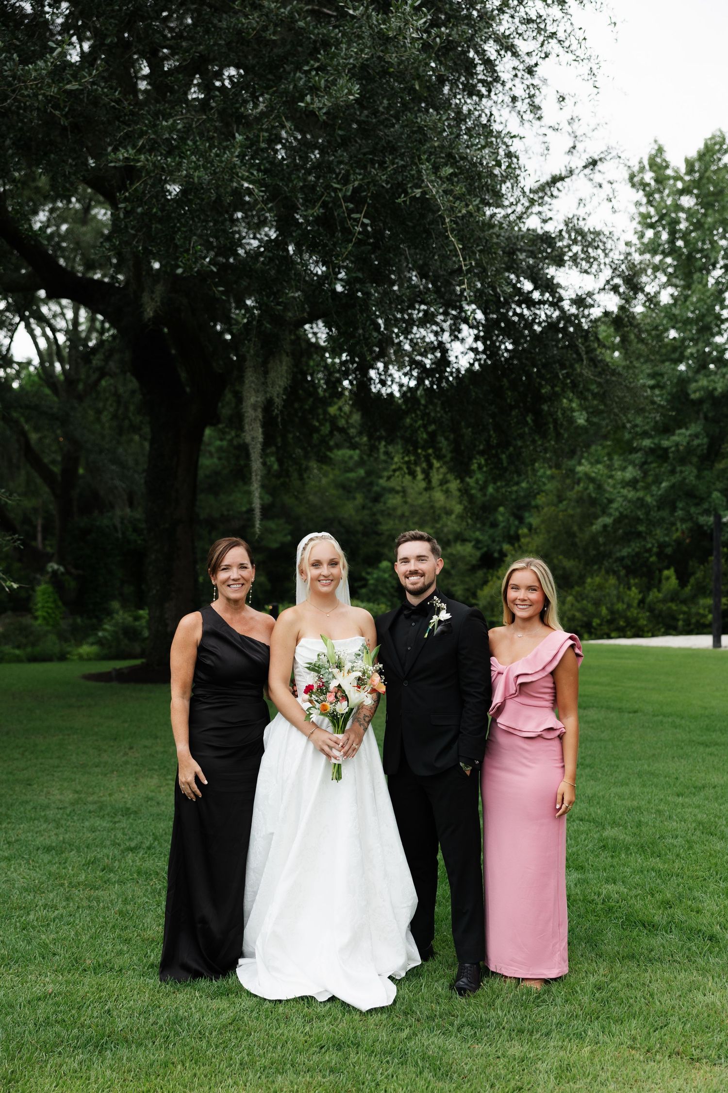 Bride and groom pose with two women in black and pink gowns on a green lawn at an outdoor wedding.