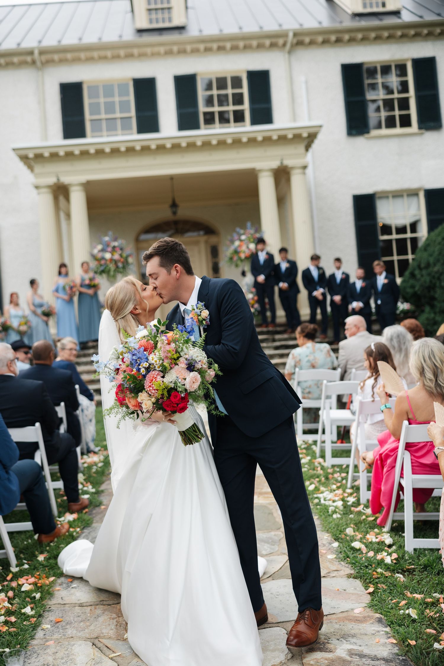 Bride and groom share a kiss at outdoor wedding ceremony in front of elegant white mansion with black shutters.
