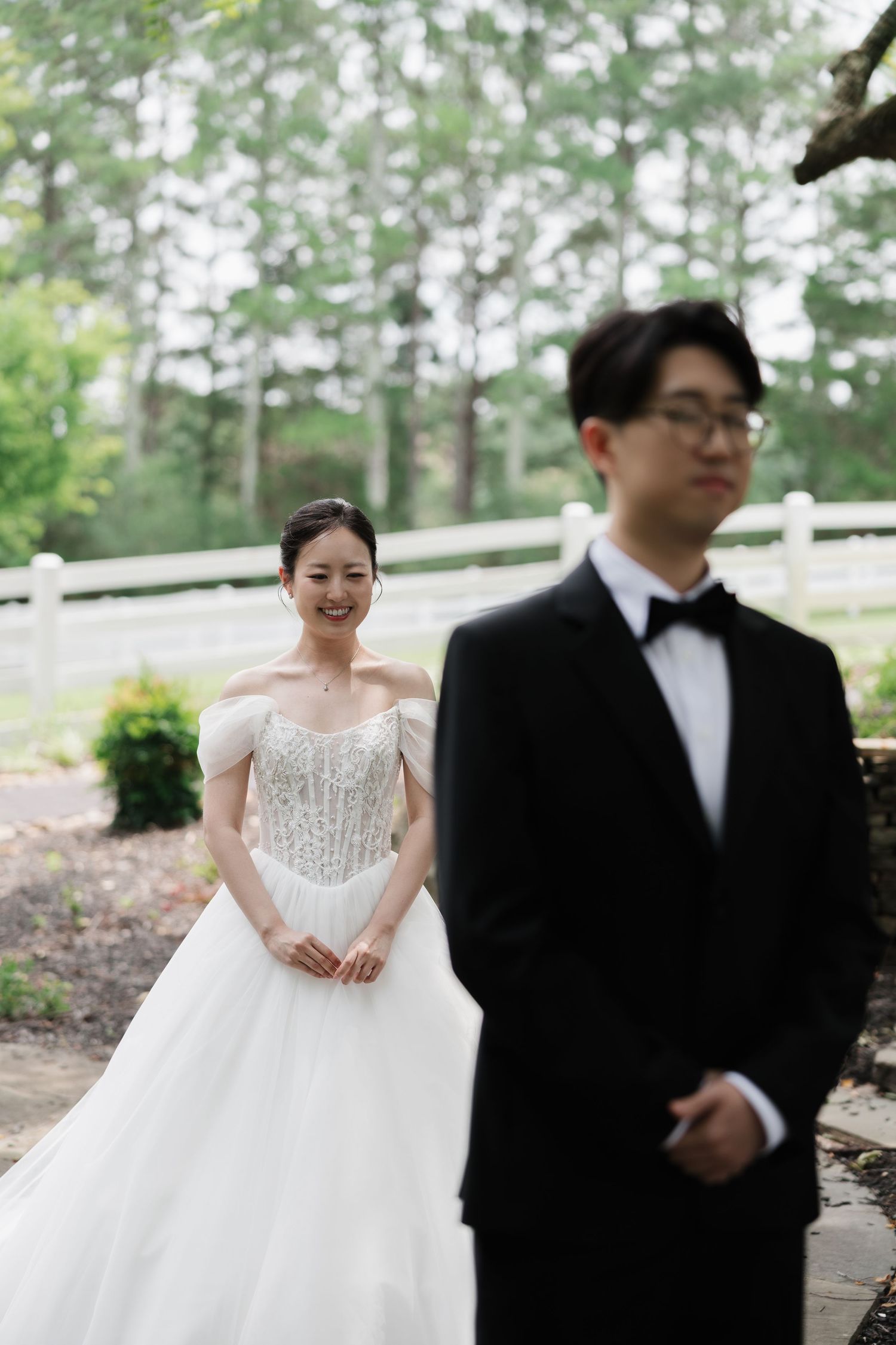 Bride in off-shoulder beaded gown smiles at groom in black tuxedo during outdoor first look wedding moment.