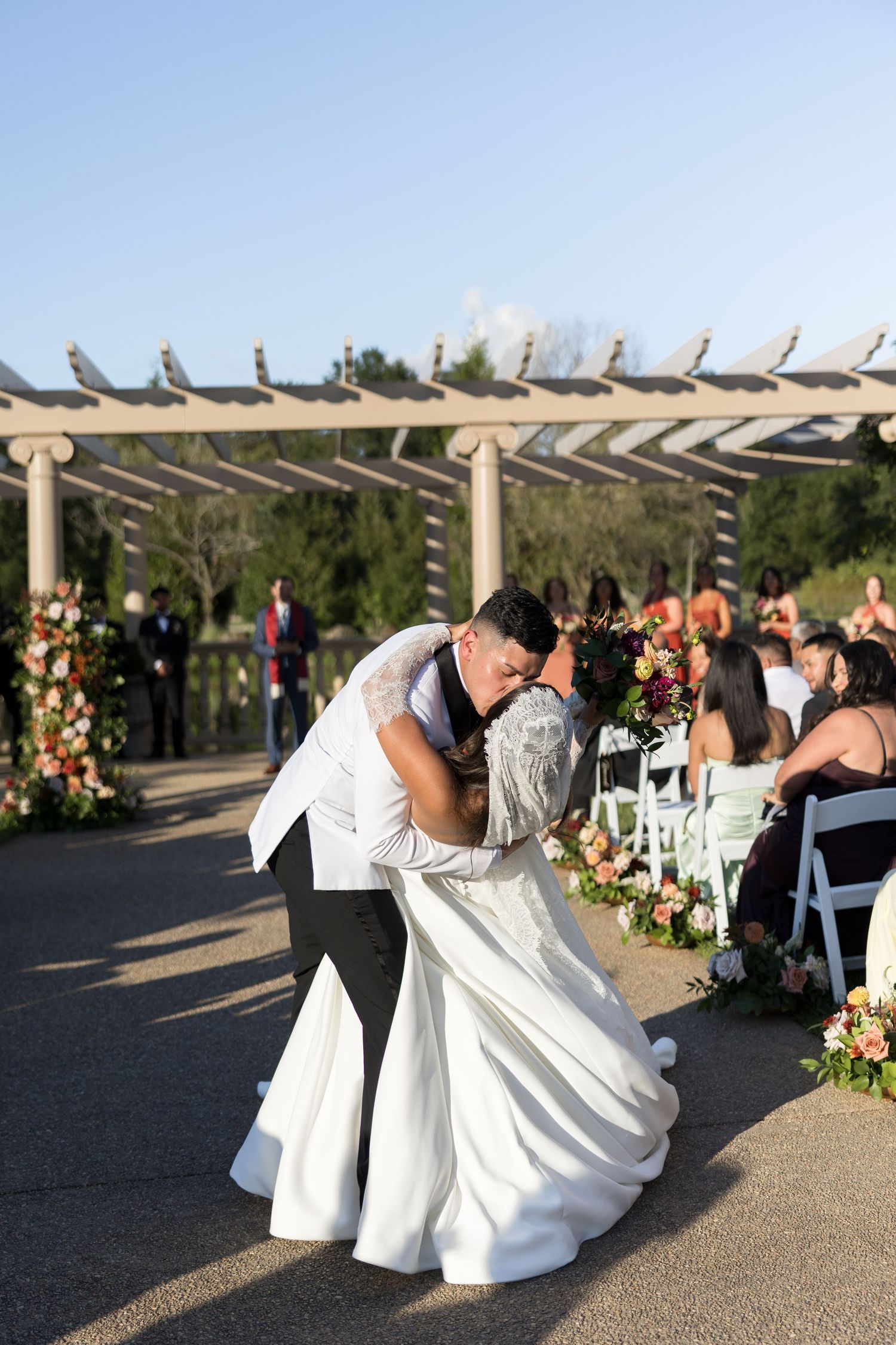 Bride and groom share a romantic dip kiss at outdoor wedding ceremony with floral arrangements and pergola backdrop.