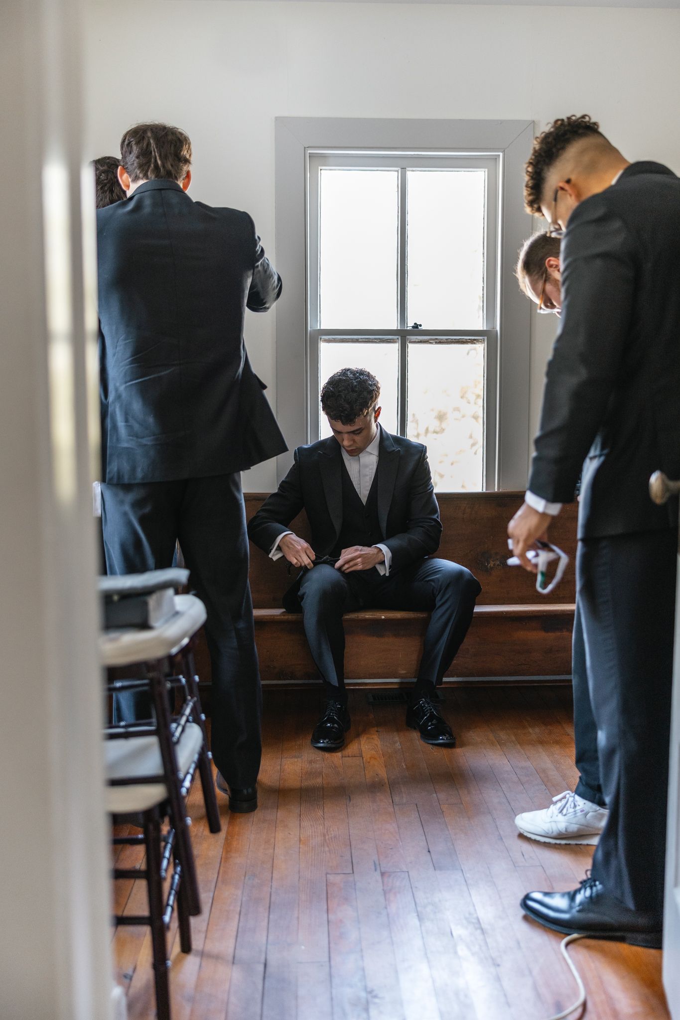 Groom and groomsmen in black suits preparing inside a rustic wooden chapel room.