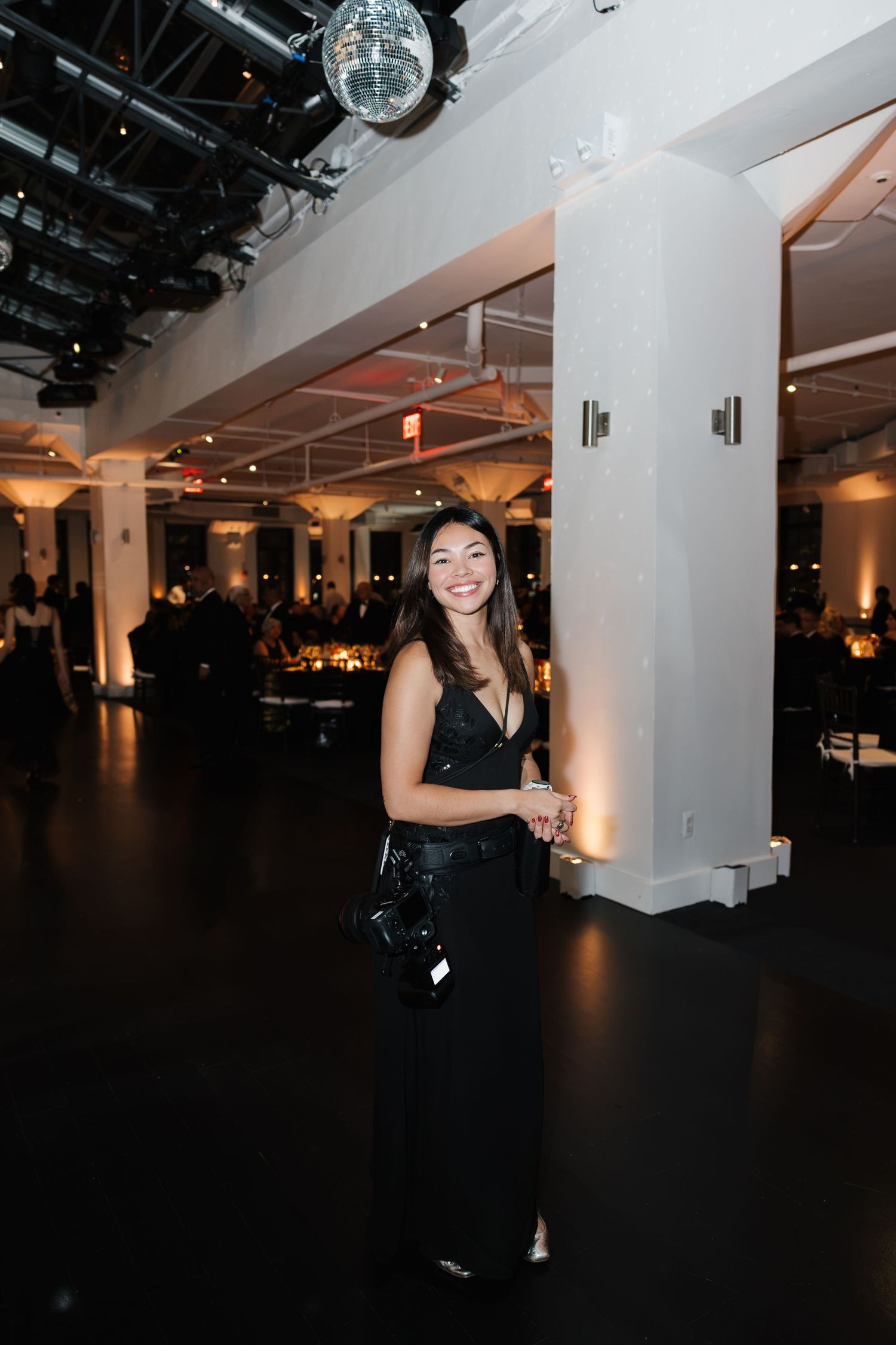 Woman in black dress smiling at elegant event venue with disco ball and ambient lighting.