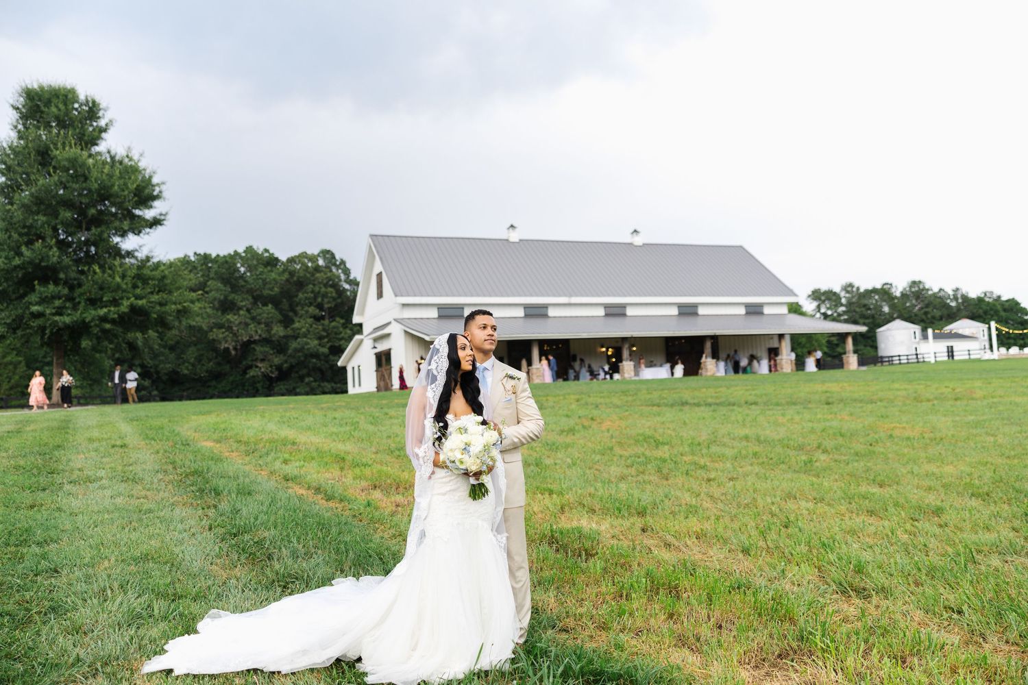 Bride and groom embrace in lush green field before a white barn wedding venue on a cloudy day.