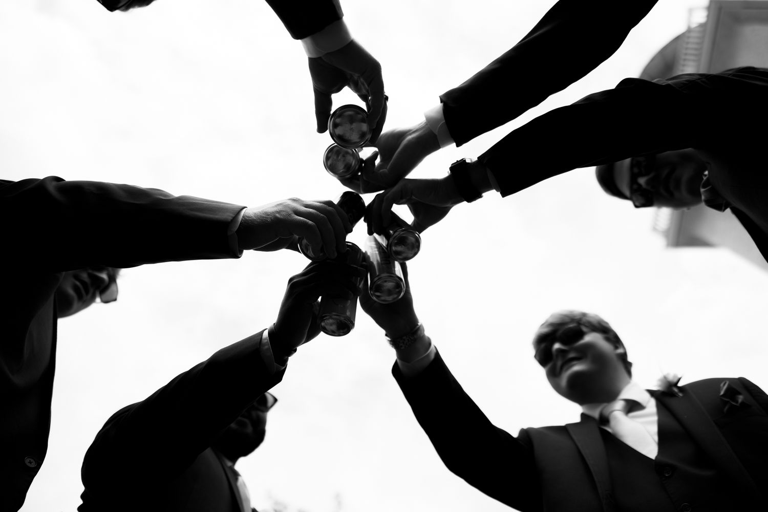 Black and white photo of men in suits toasting drinks together in a circle, viewed from below.