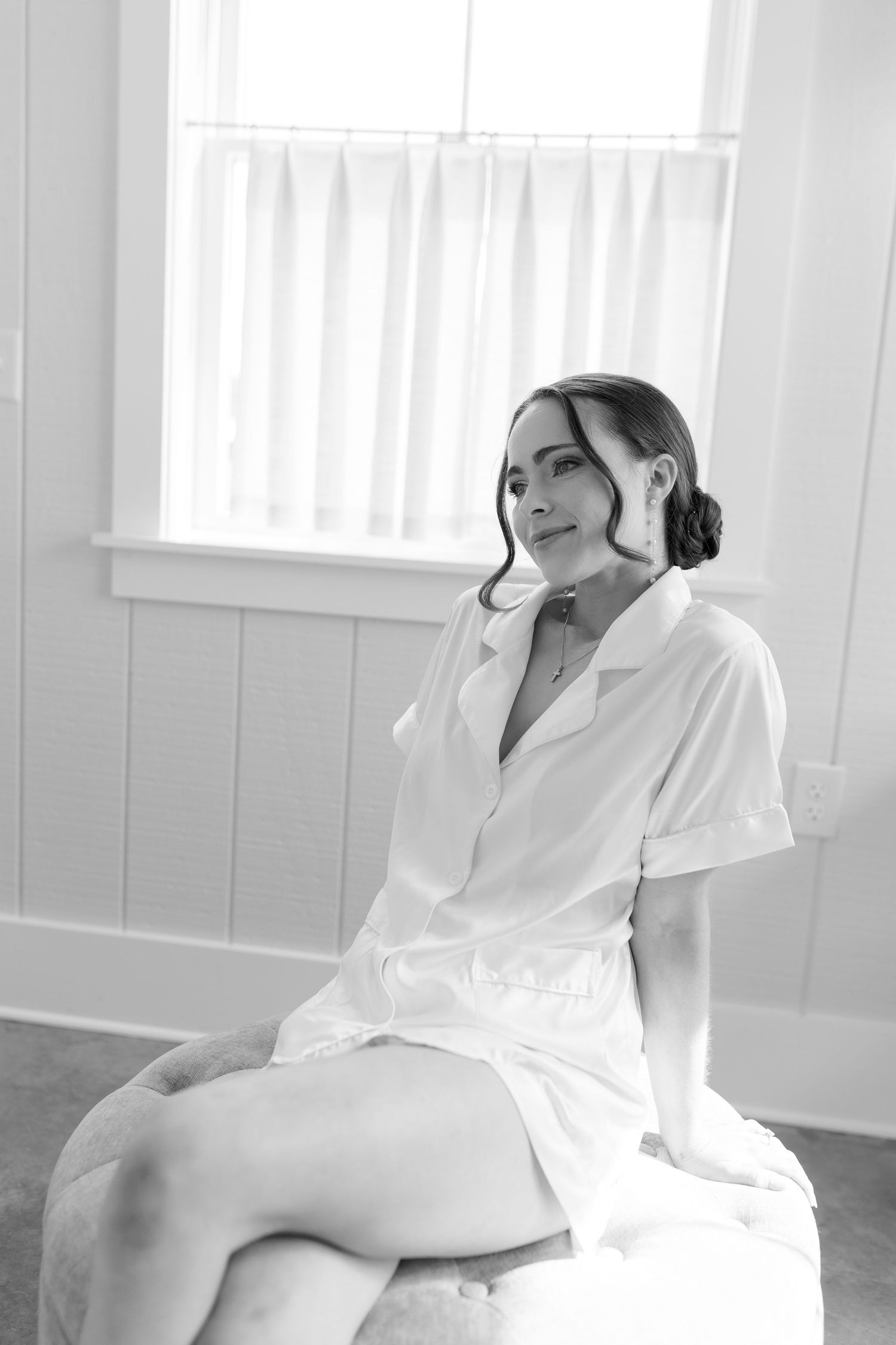 Black and white portrait of bride in white robe sitting thoughtfully by bright window.