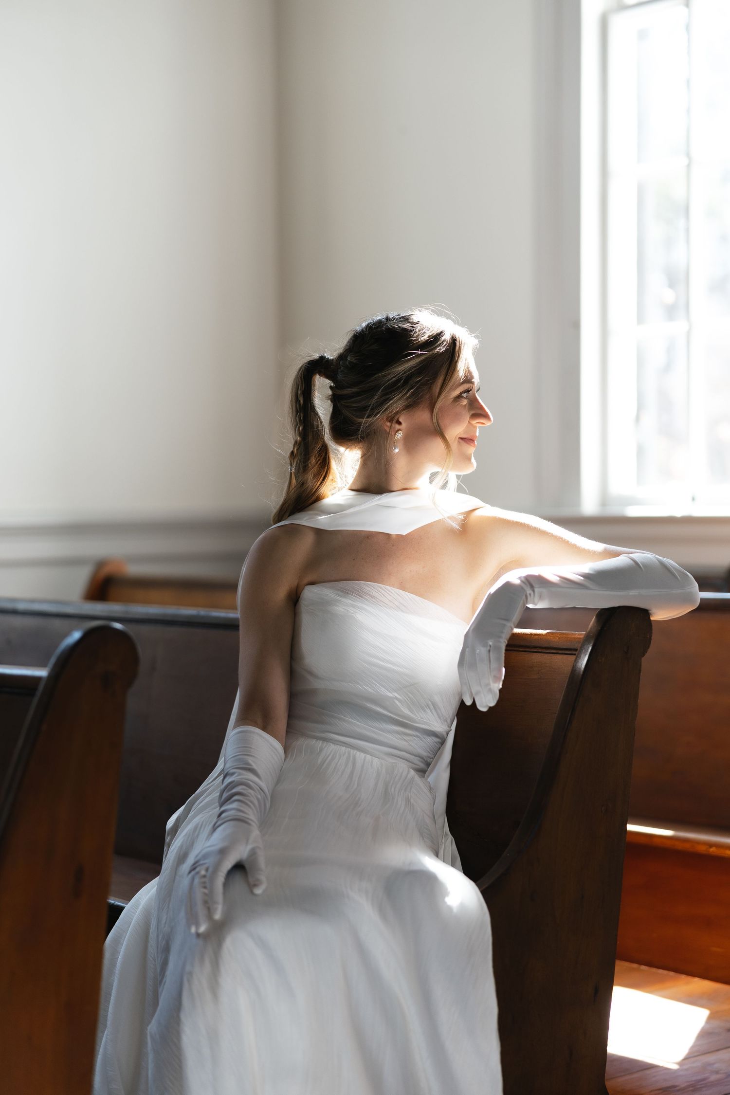 Bride in white gown and gloves sitting on church pew gazing out a sunlit window.