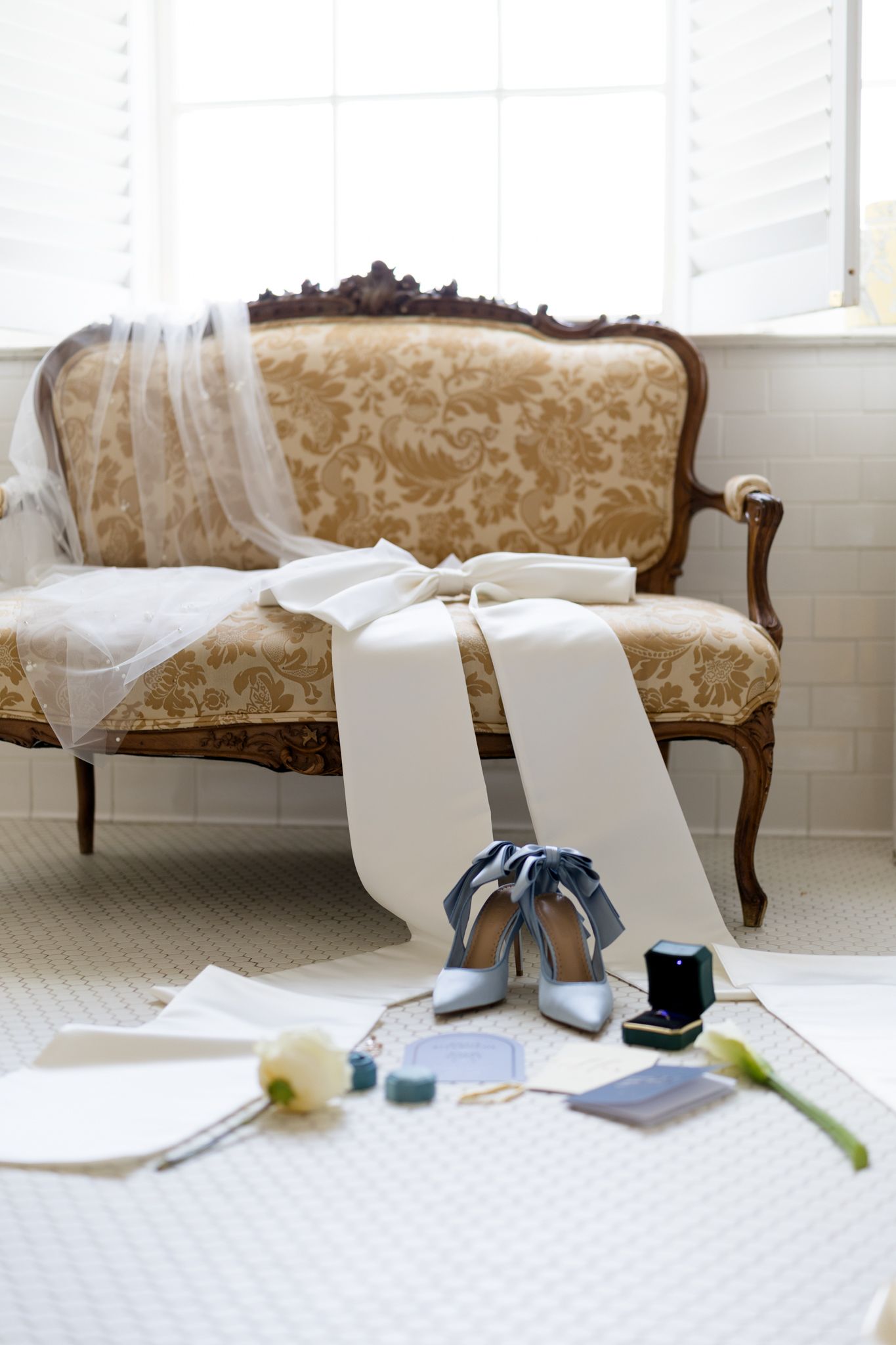 Blue satin heels and ring box on floor before vintage gold brocade settee with wedding veil draped over it.