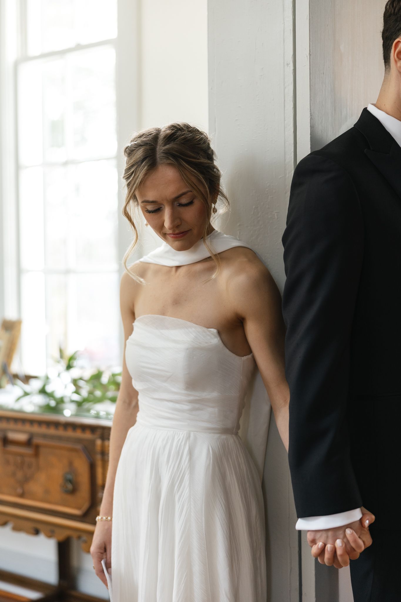 Elegant bride in white halter wedding dress holds grooms hand while gazing down by bright window.