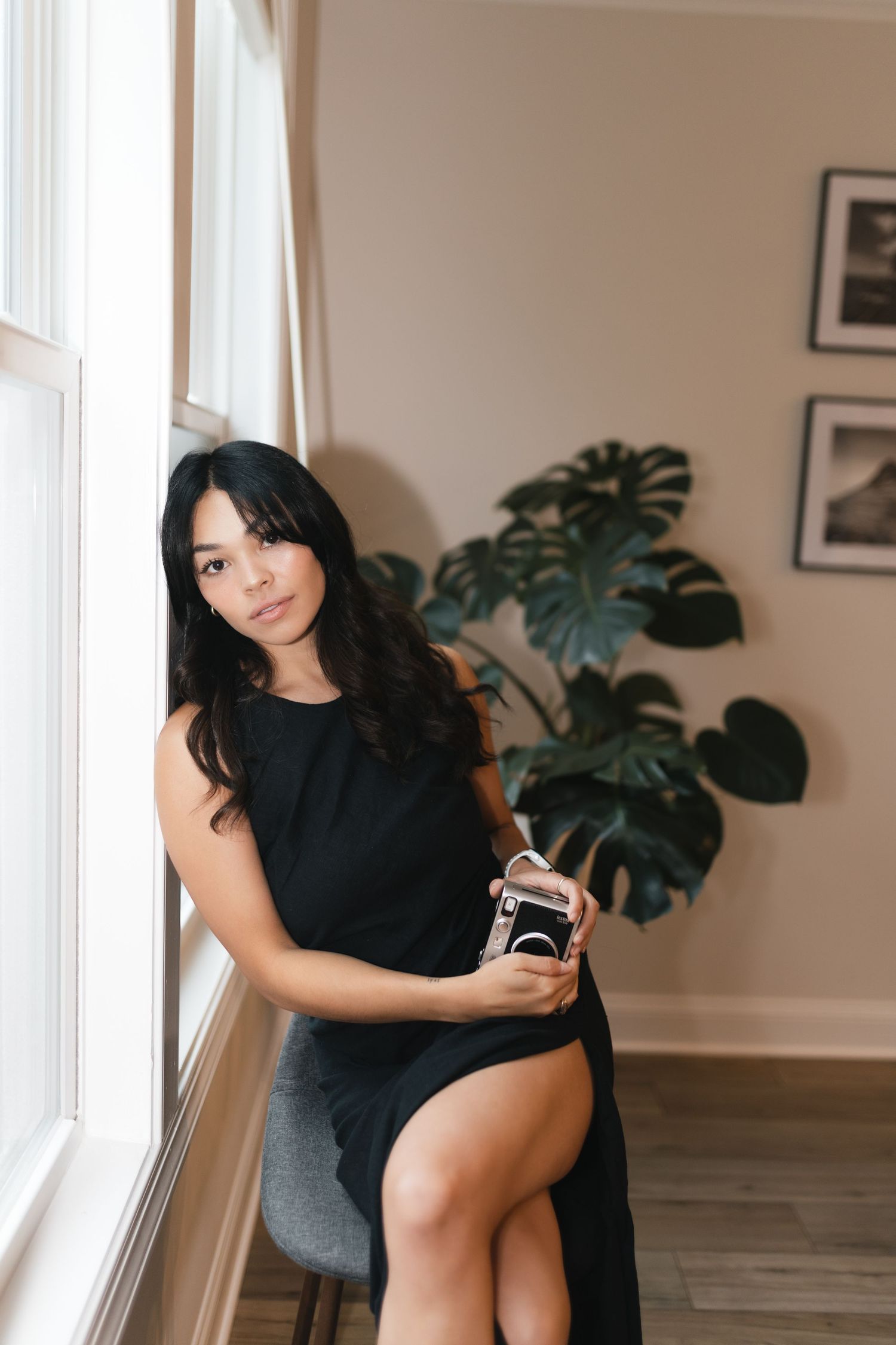 Woman in black dress with slit sits by window holding vintage camera with monstera plant behind.