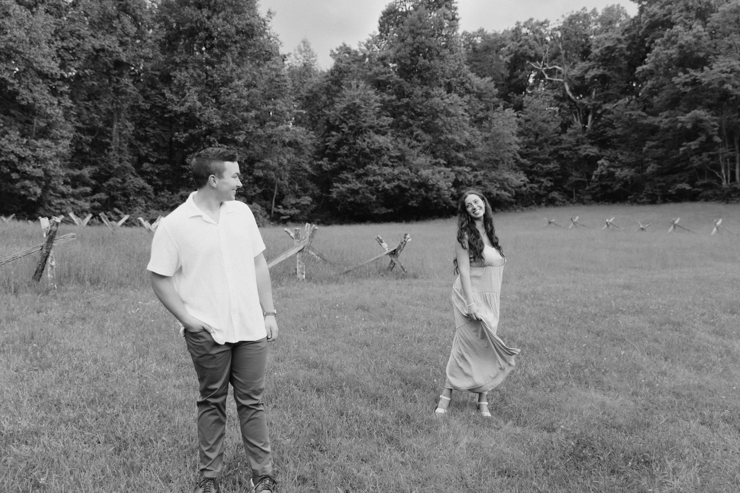 A couple shares a playful moment in a black and white photo on a grassy meadow surrounded by trees.