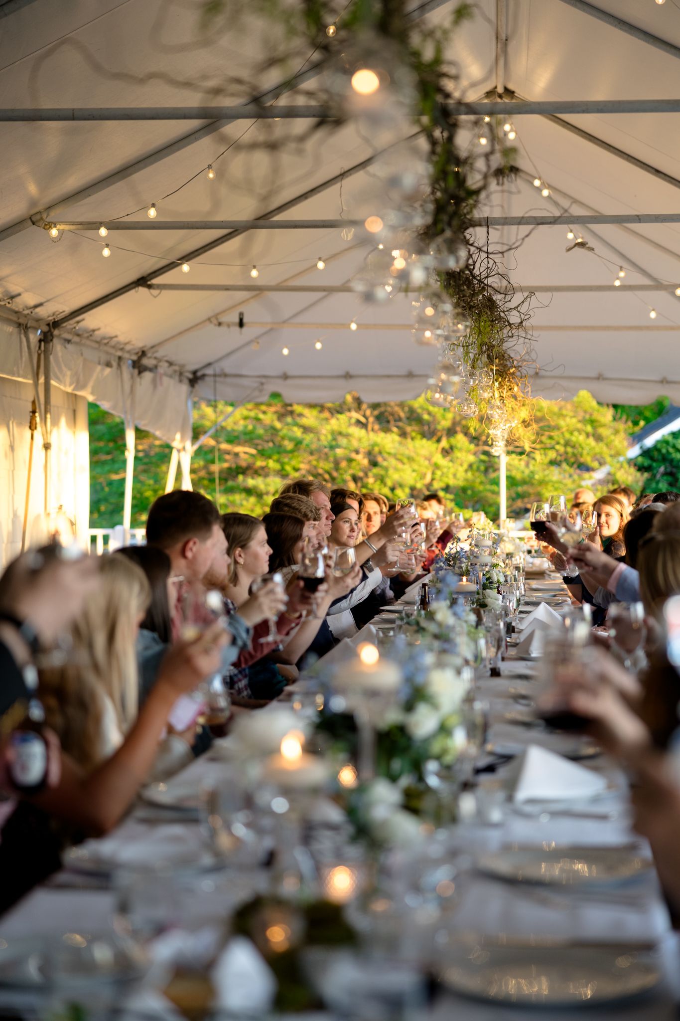 Guests raise glasses in a toast at a long elegantly decorated rehearsal dinner table under a white tent with string lights.