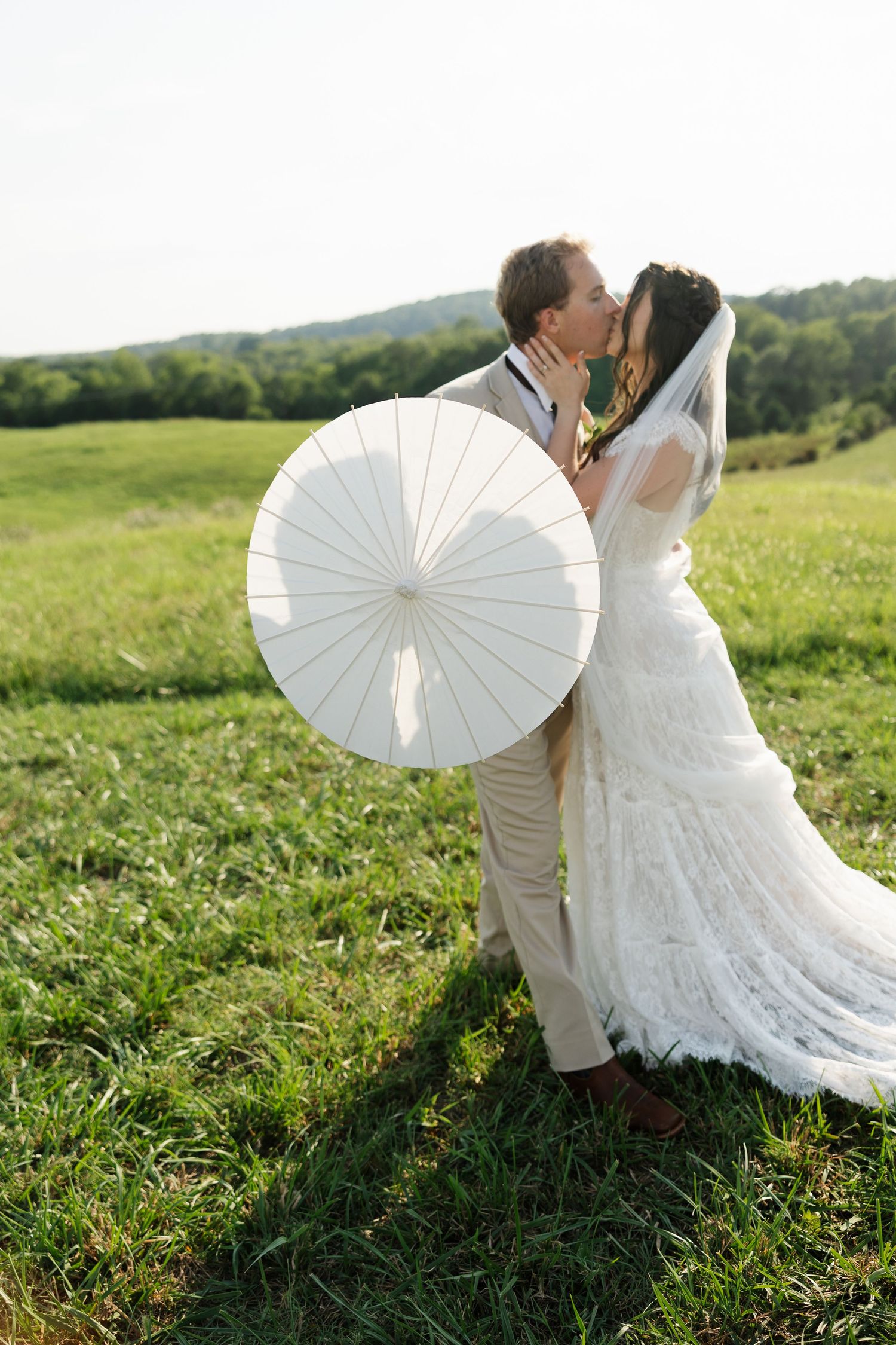 Bride and groom kissing in green field, bride holding white parasol, wearing lace gown with veil, groom in tan suit.