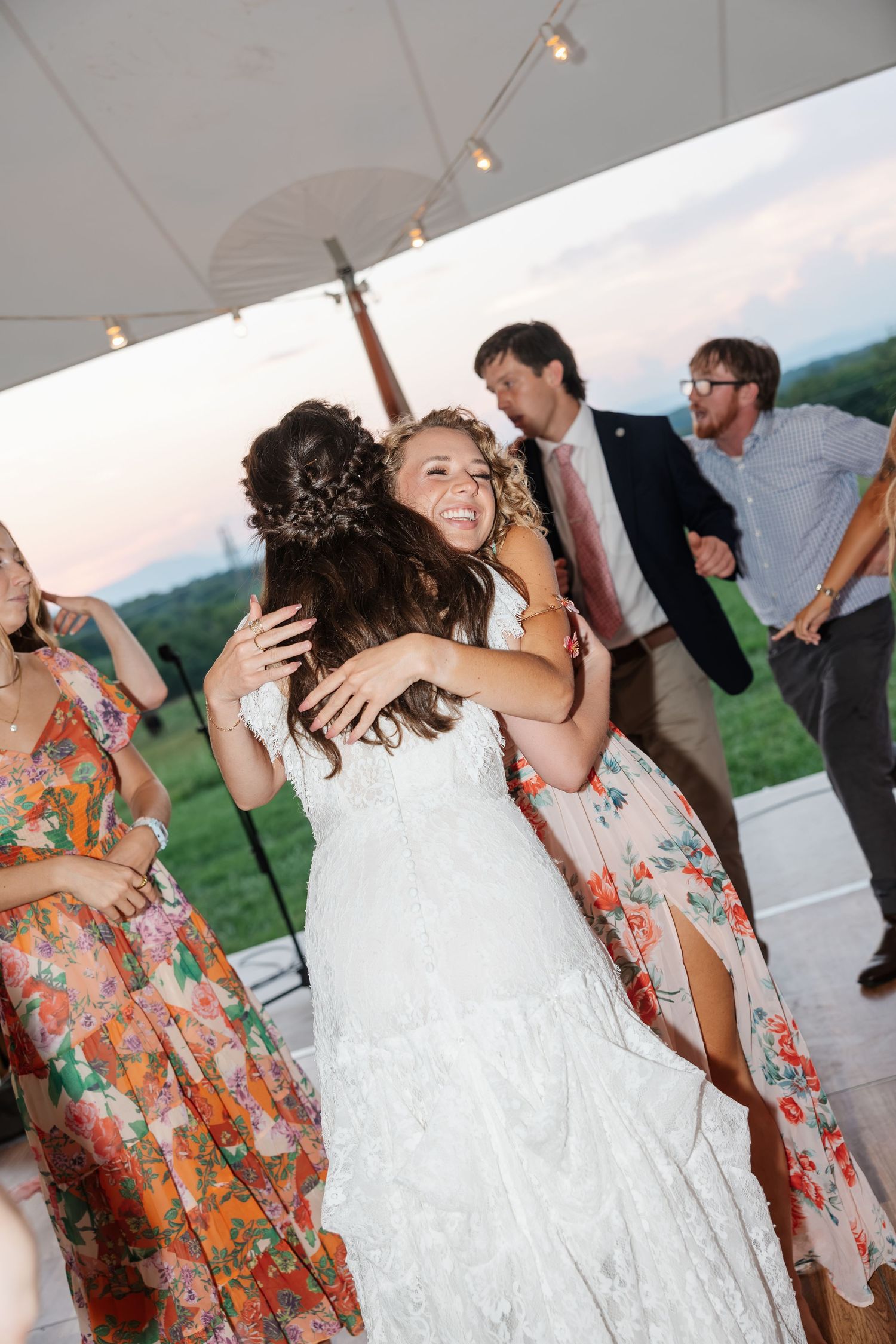 Bride in white lace gown joyfully hugging a guest on the dance floor at outdoor wedding reception.