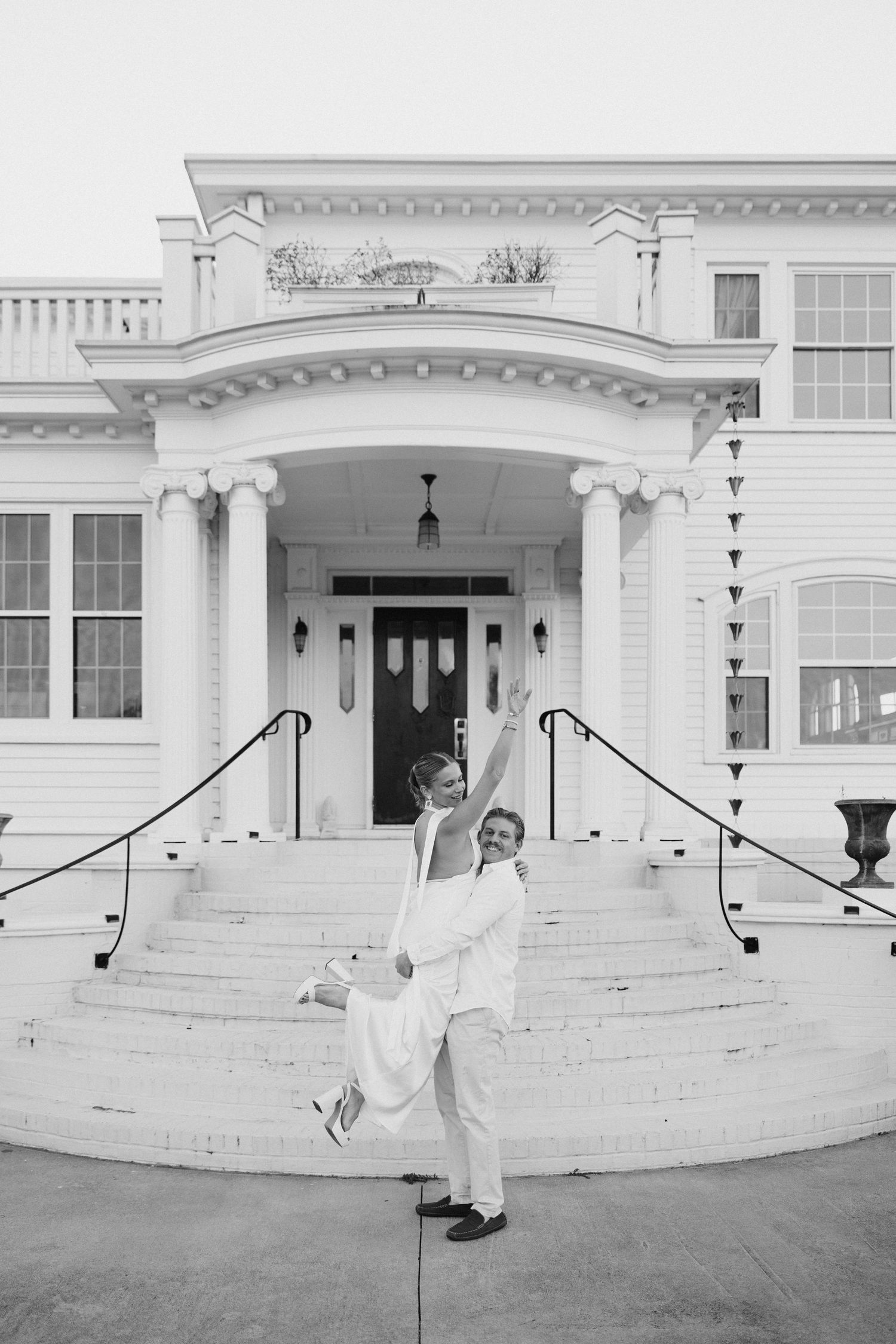 Joyful couple in white wedding attire posing on grand mansion steps in black and white photo.