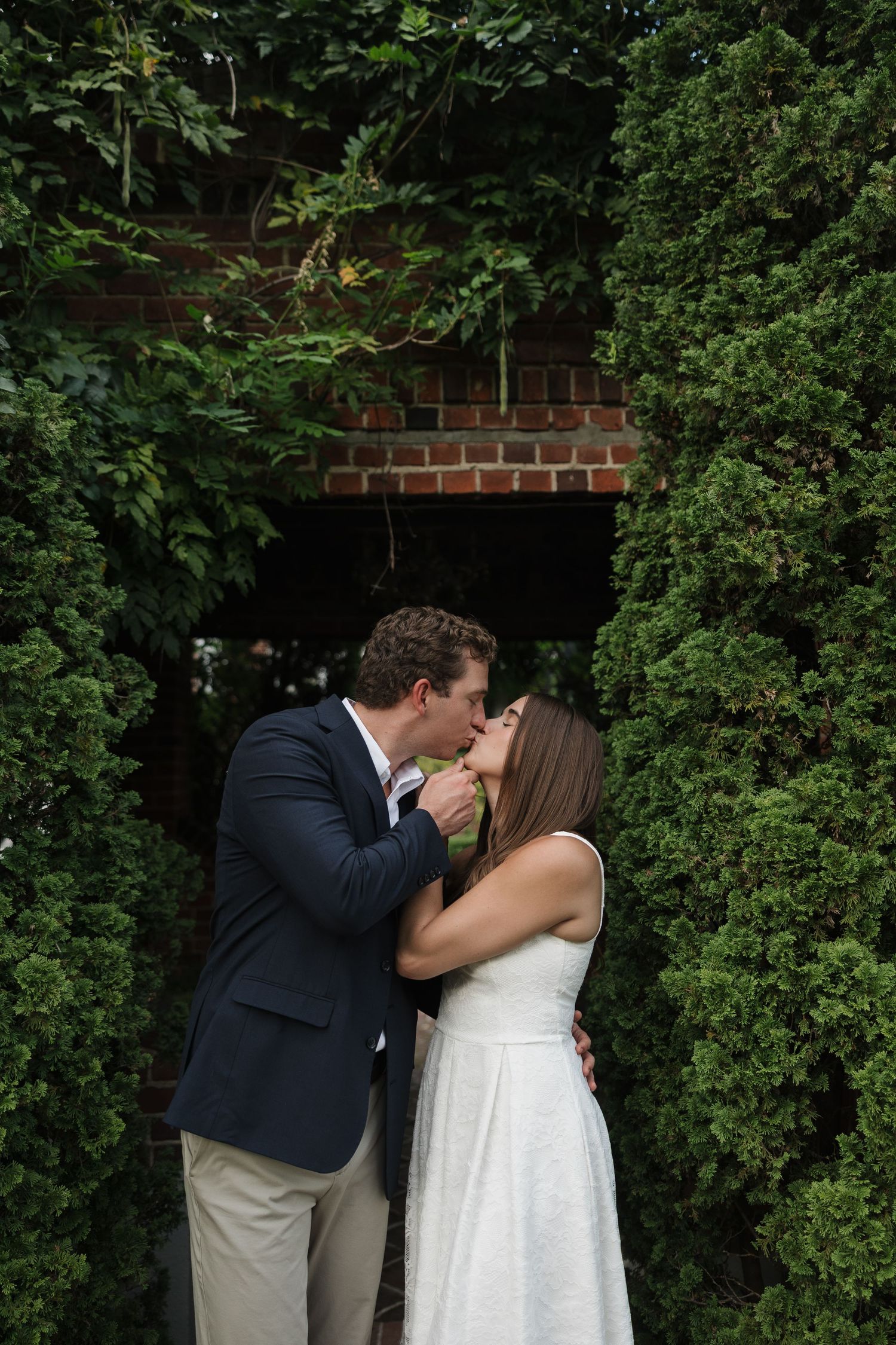 Couple sharing a romantic kiss in a lush garden archway, bride in white dress and groom in navy suit.