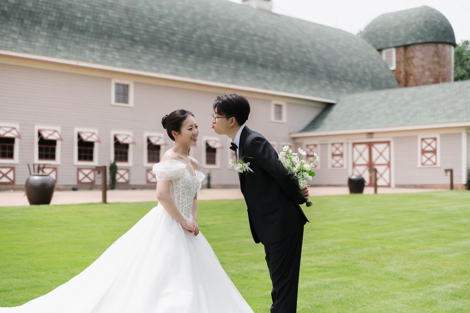 Bride and groom share a romantic moment on lush green lawn outside a charming barn wedding venue.