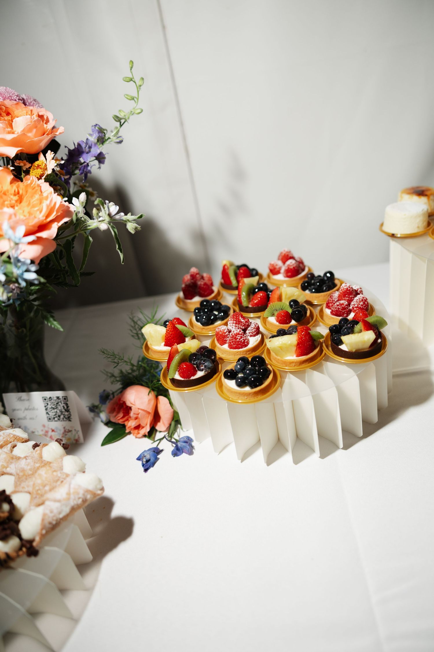 Elegant dessert table with fruit tarts topped with fresh berries on a white display stand, surrounded by colorful floral arrangements.