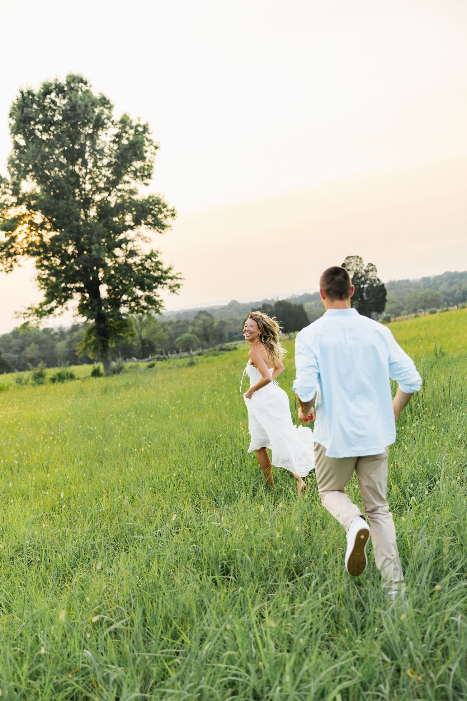 A couple holding hands running through a lush green meadow at sunset, woman in white dress, man in light blue shirt.
