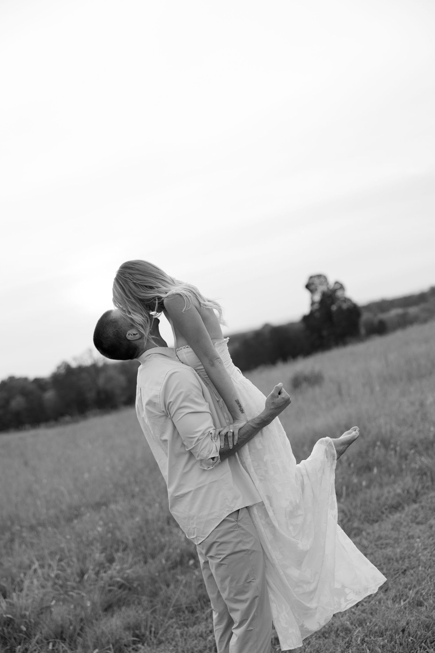 Black and white photo of groom lifting bride in a romantic field during outdoor engagement session.
