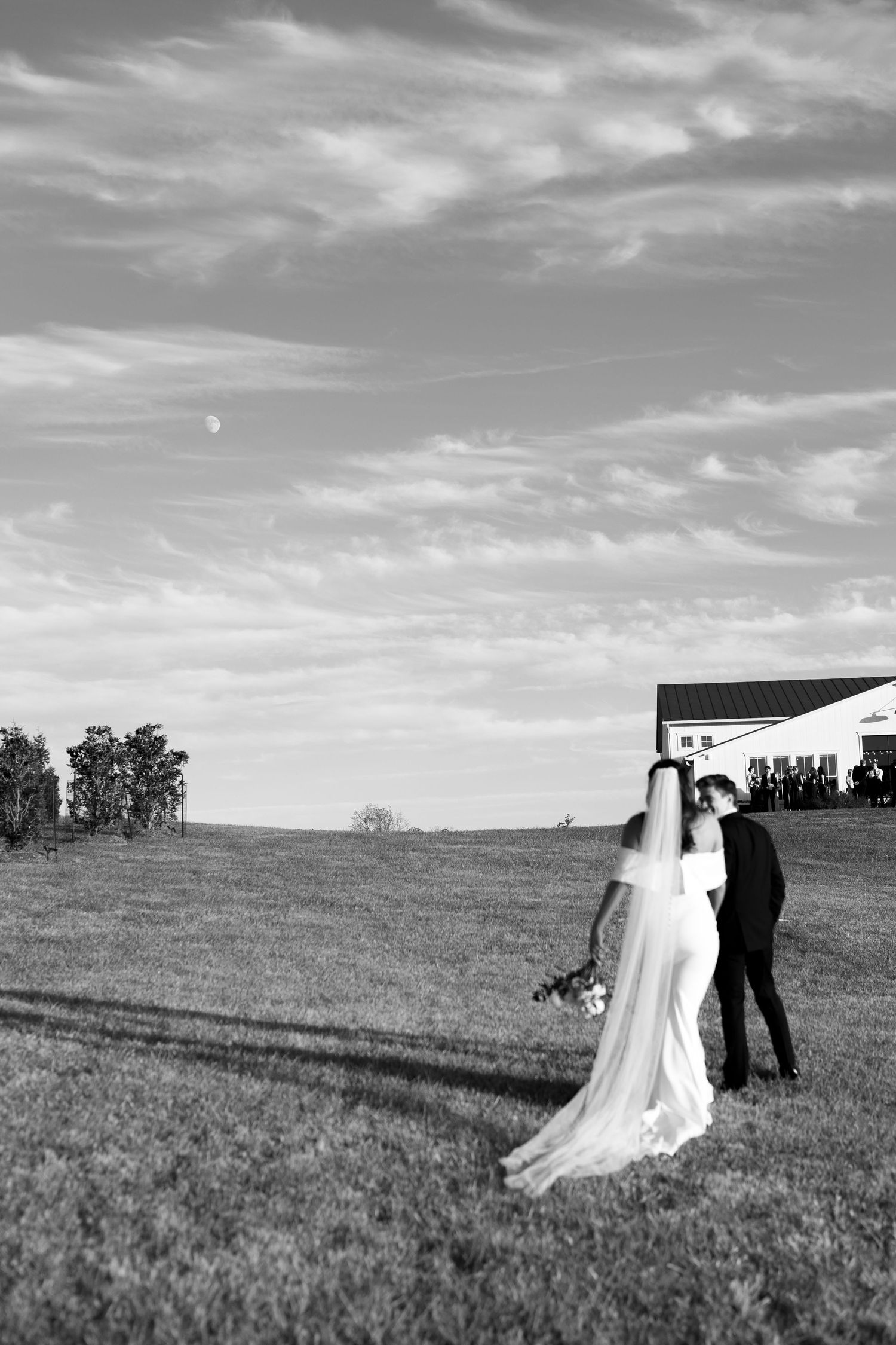 Black and white photo of bride and groom walking across open field toward white barn under cloudy sky.