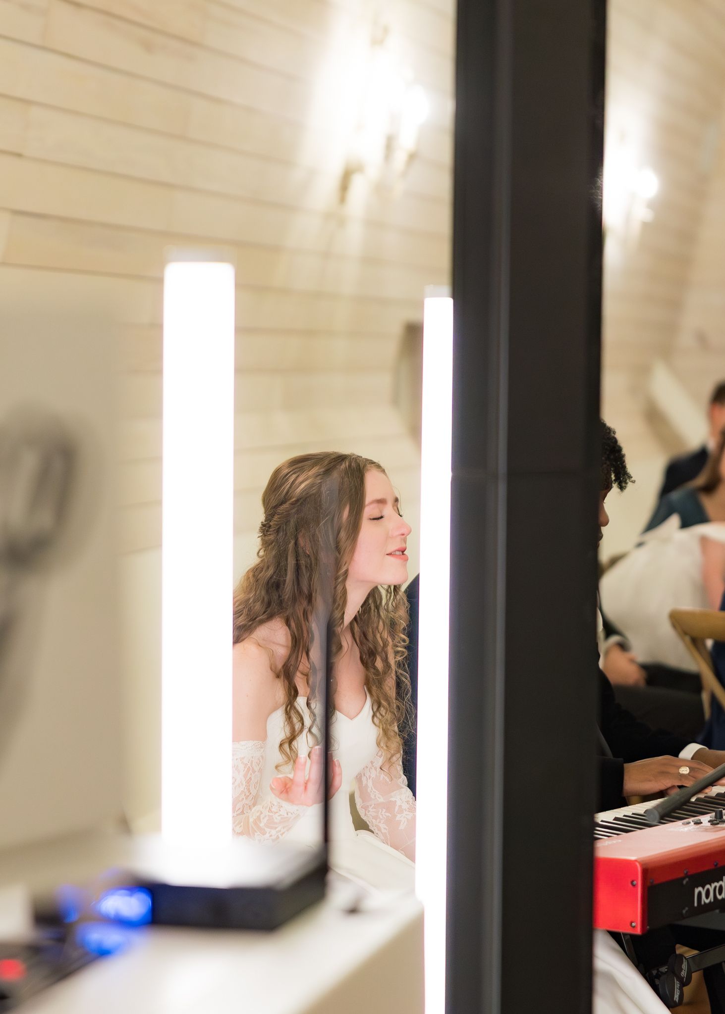 Bride with curly hair smiles while getting ready, reflected in lighted vanity mirrors backstage.