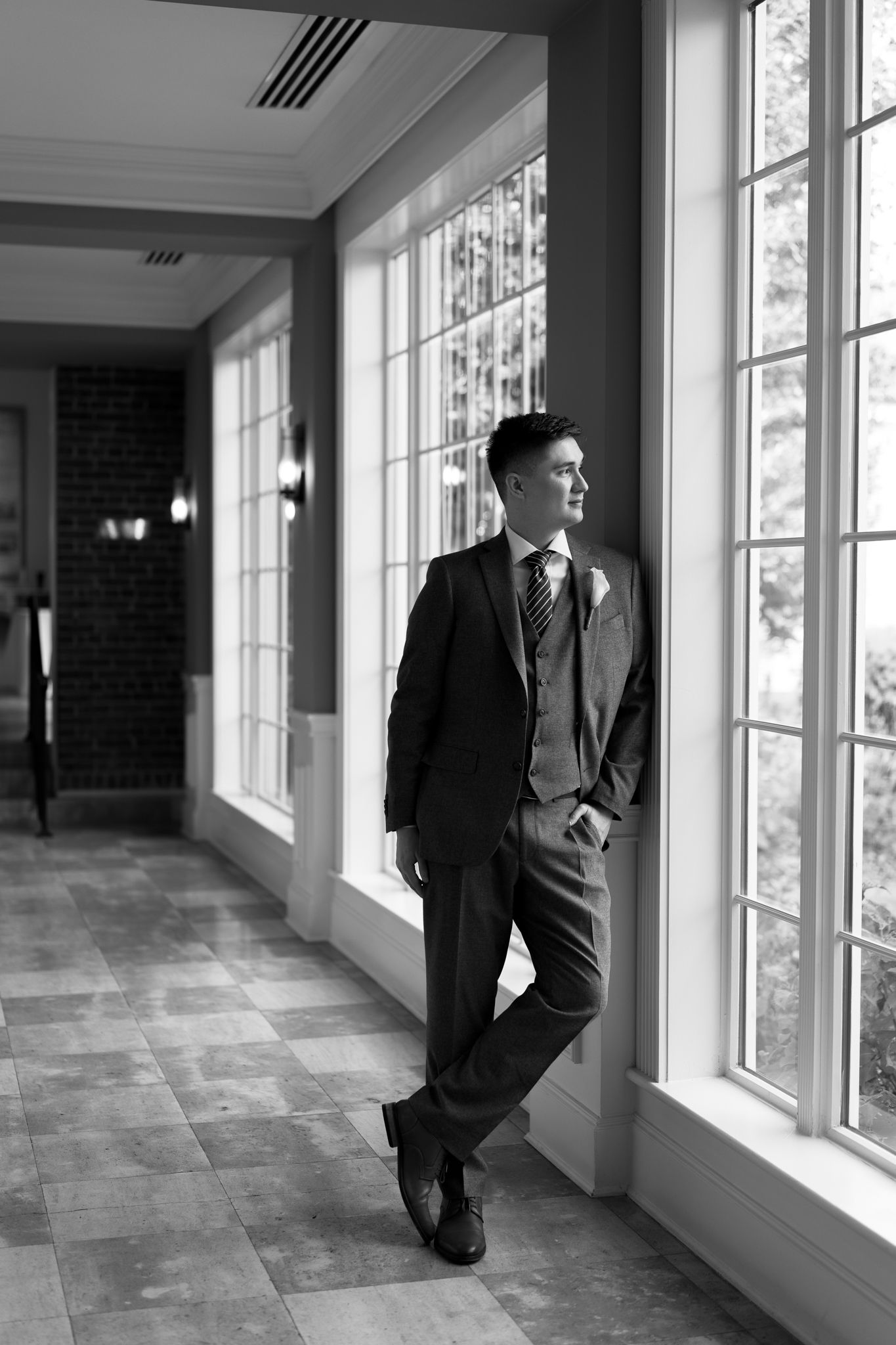 Groom in dark suit leaning against window in elegant hallway, black and white wedding portrait.