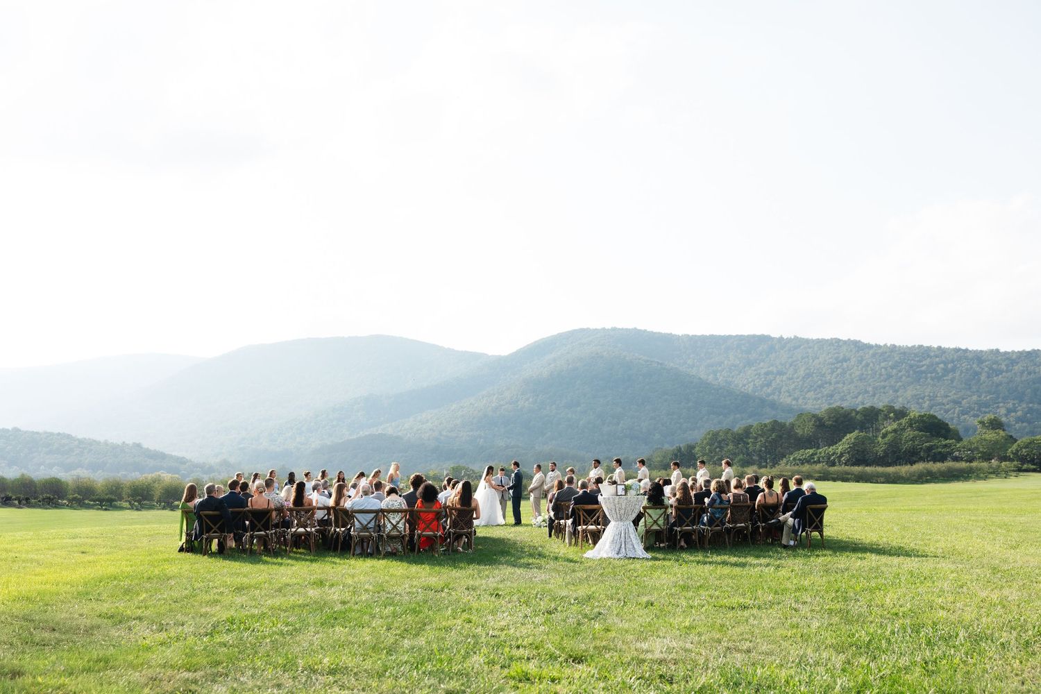 Outdoor wedding ceremony in a green meadow with mountain views, guests seated in rows facing the couple on a sunny day.