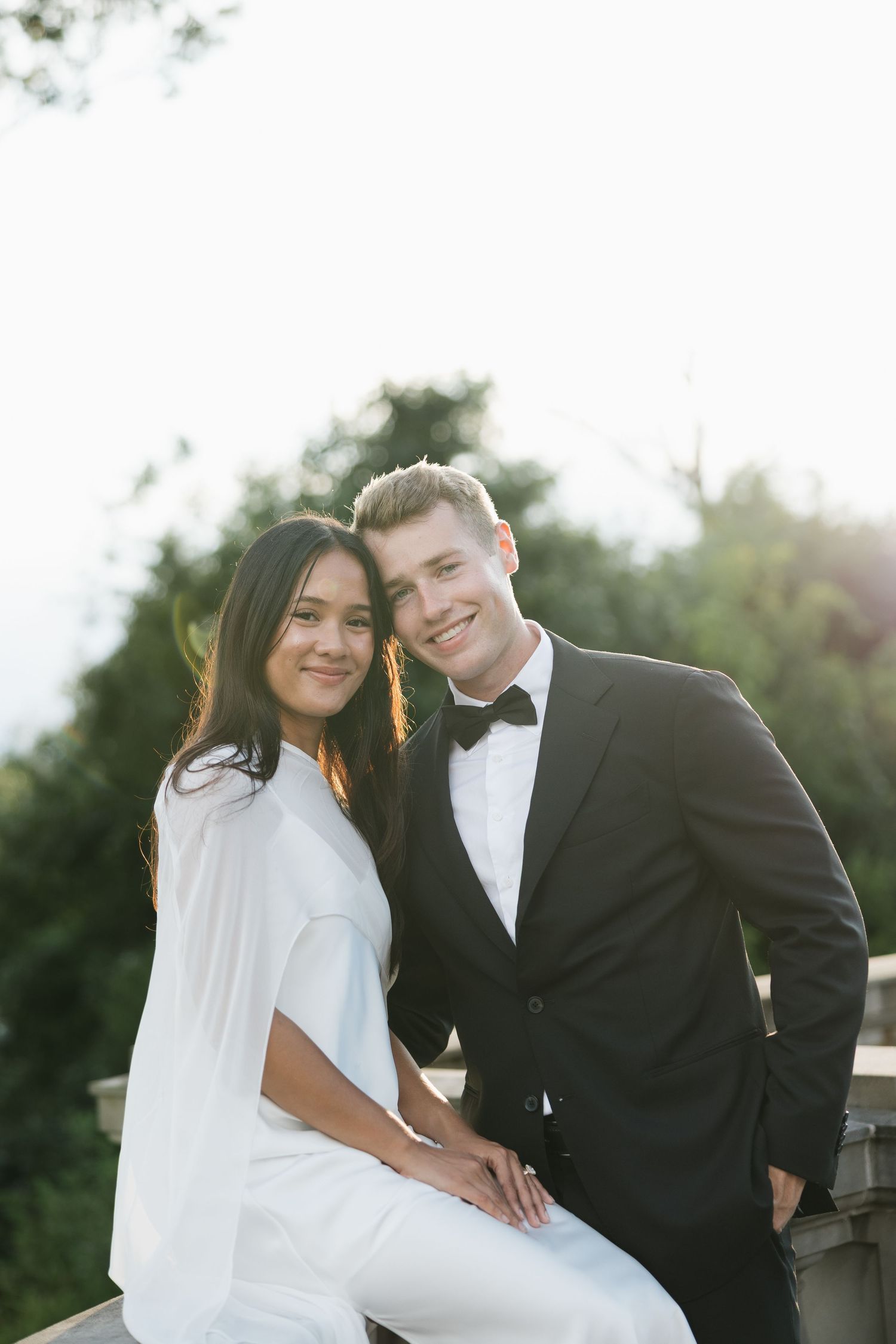Elegant couple in formal attire posing outdoors, man in black tuxedo and woman in white outfit, smiling at camera.