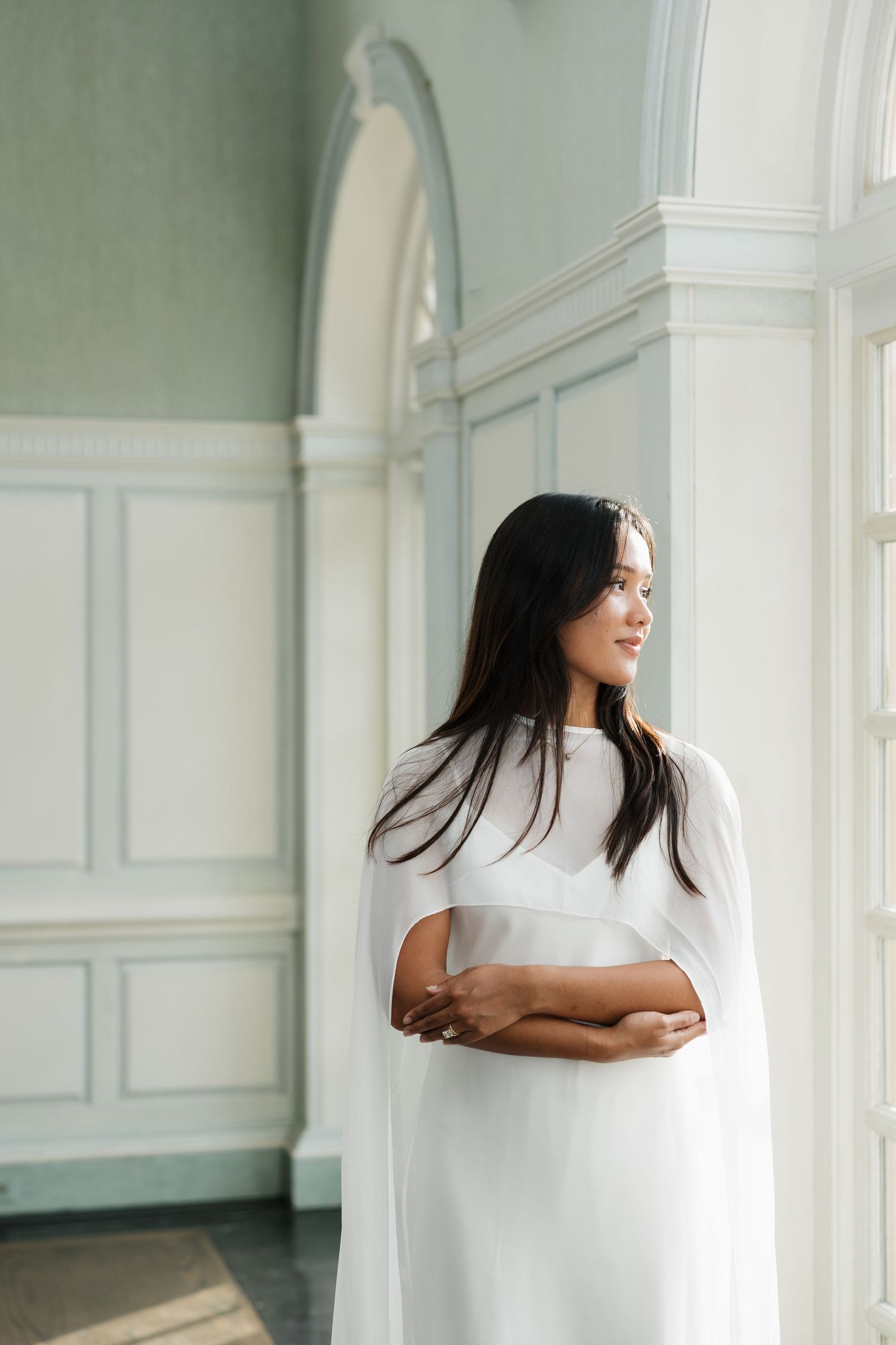 Woman in white cape dress stands by ornate window in elegant room, looking sideways with arms crossed.