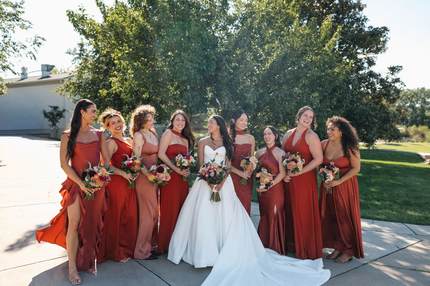 Bride in white gown poses with bridesmaids in red dresses holding bouquets outdoors on sunny day.