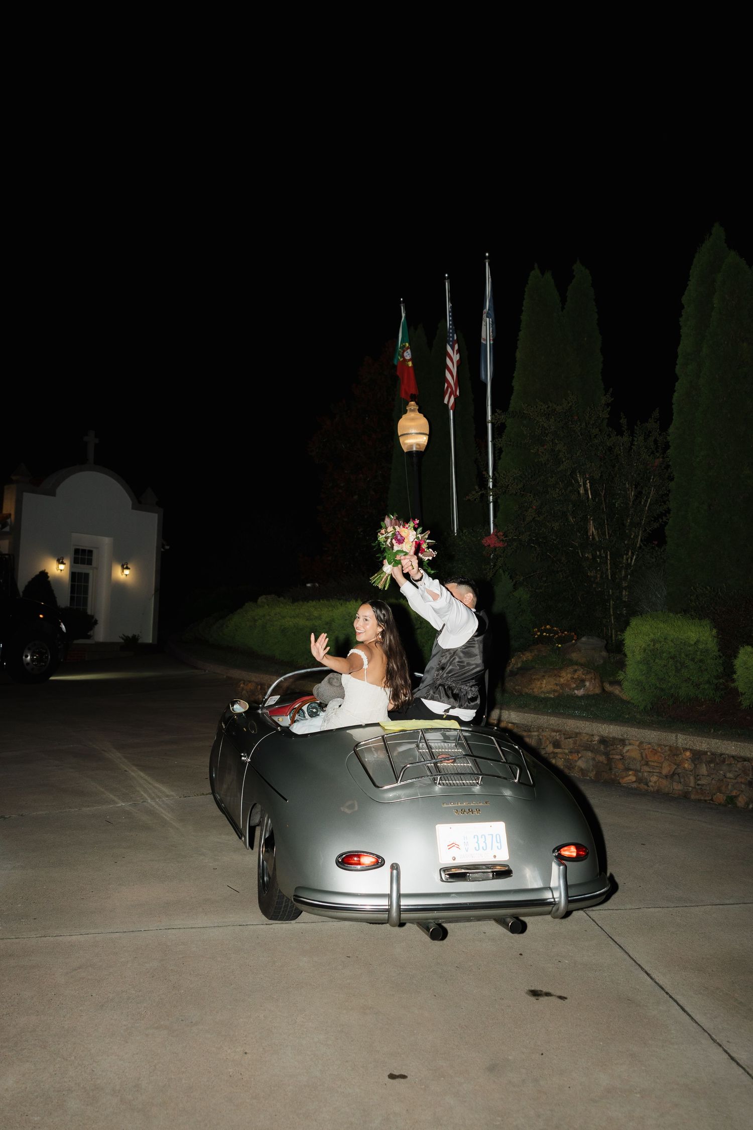 Newlyweds wave from a vintage silver sports car at night, bride in white gown with floral crown, groom in suit.