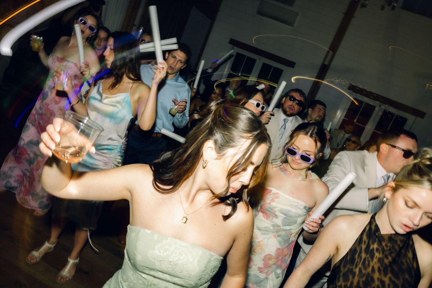 Energetic wedding reception dance floor filled with guests in colorful dresses holding light-up foam sticks.