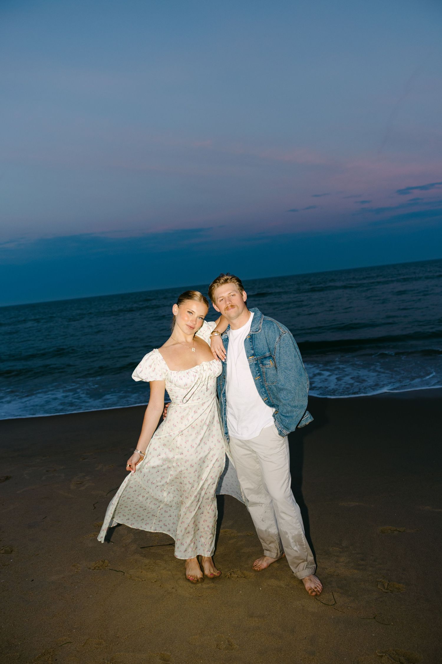 Couple poses together smiling on sandy beach during a beautiful blue and pink twilight sunset.