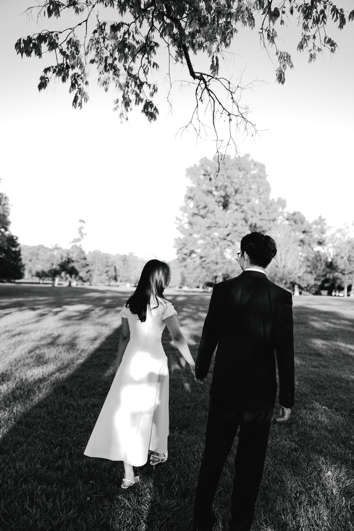 Black and white photo of a bride and groom holding hands walking through a lush green park.