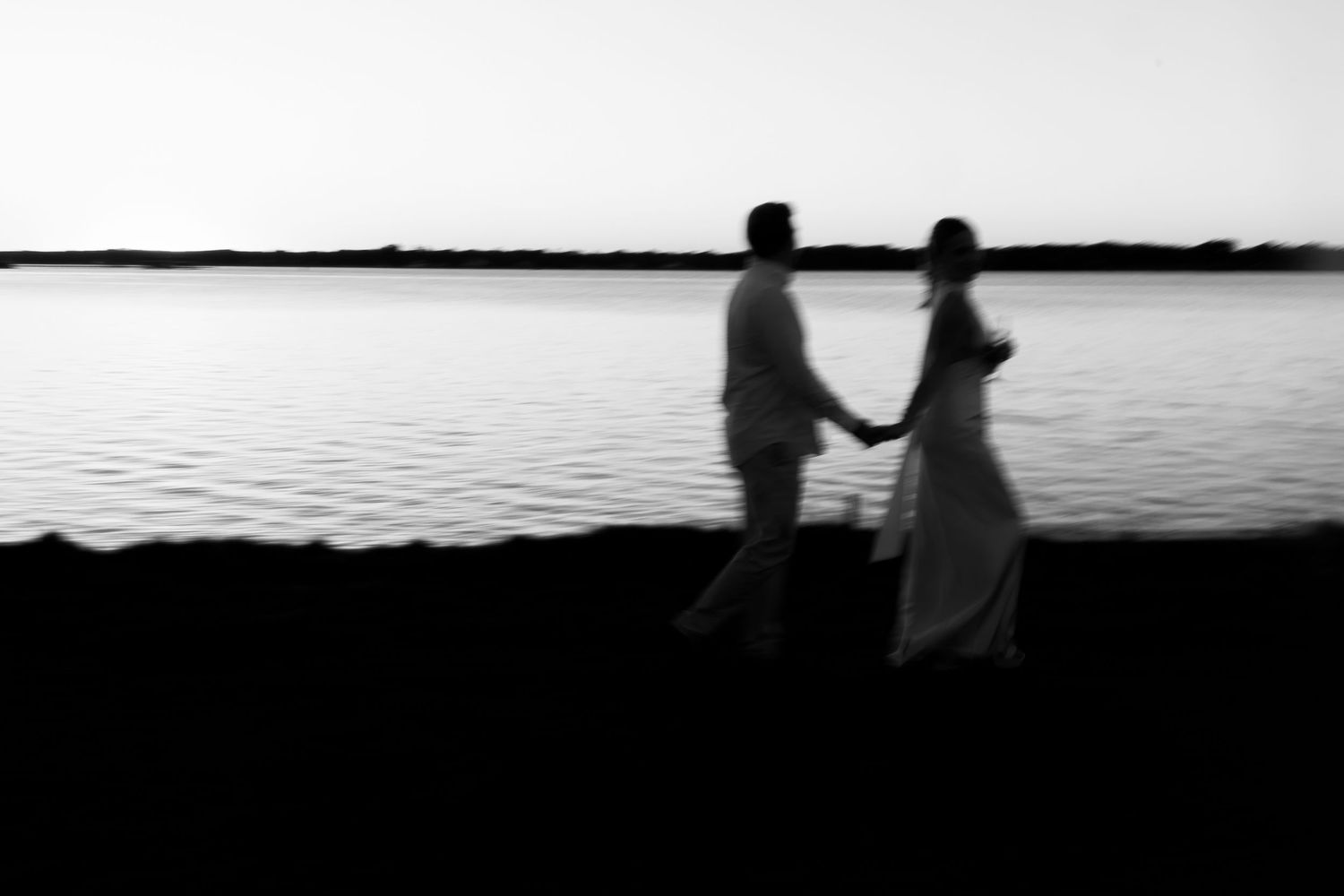 Blurry black and white photo of a couple holding hands by a calm lake at dusk.