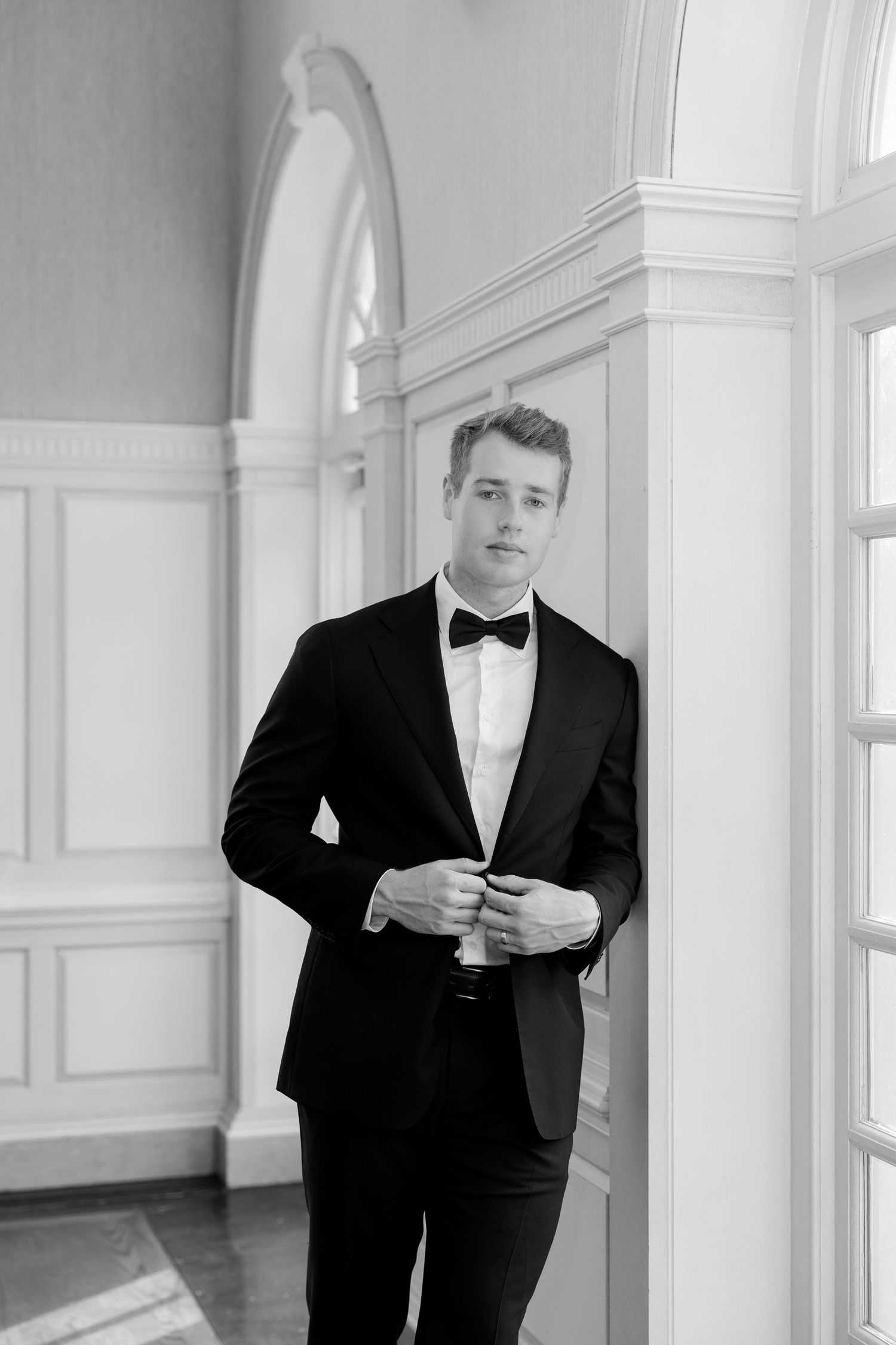 Black and white portrait of a young man in a tuxedo with bow tie leaning against a pillar in an elegant hall.