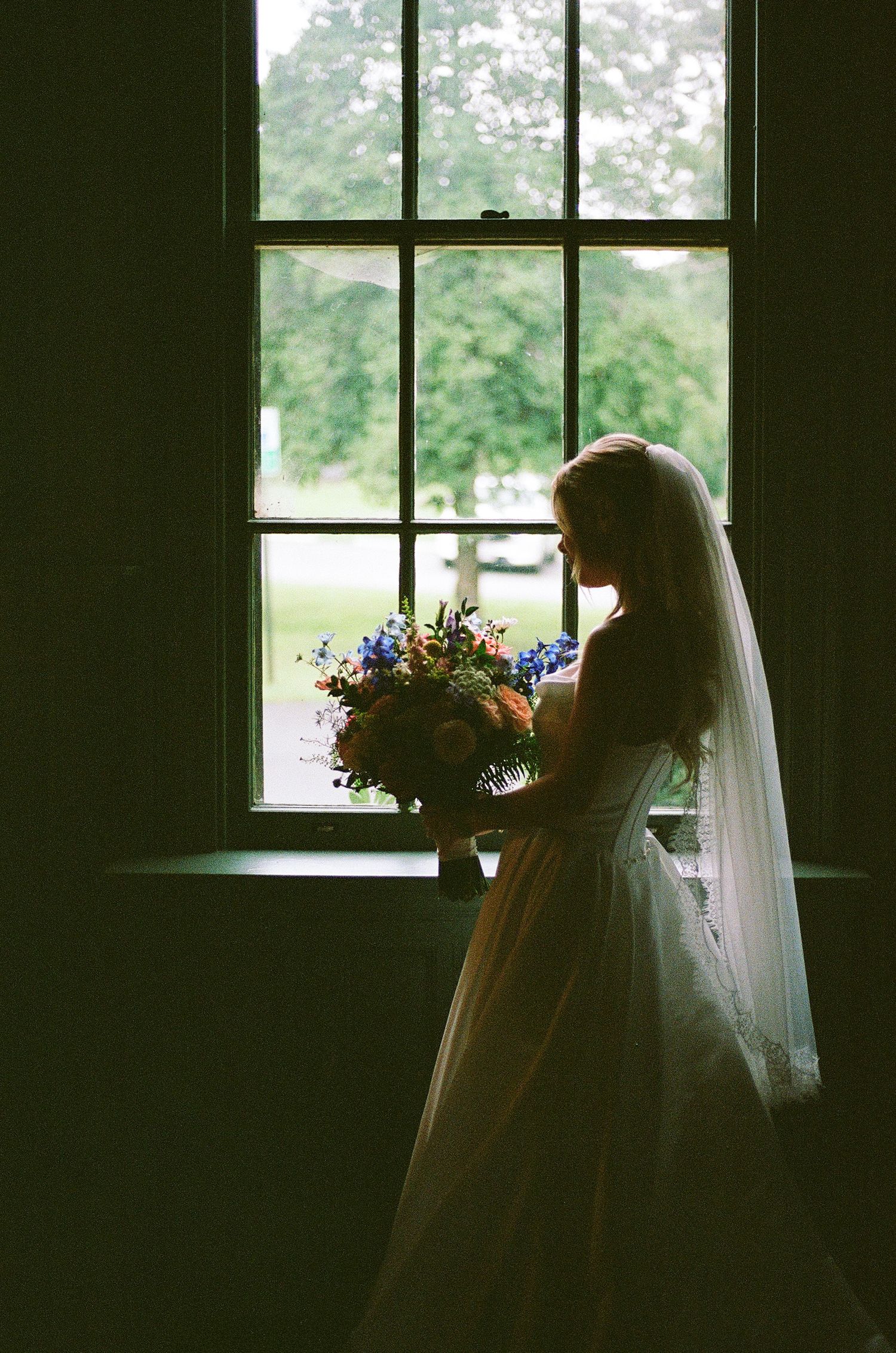 Bride in white gown and veil holds colorful bouquet by sunlit window, elegant wedding portrait.