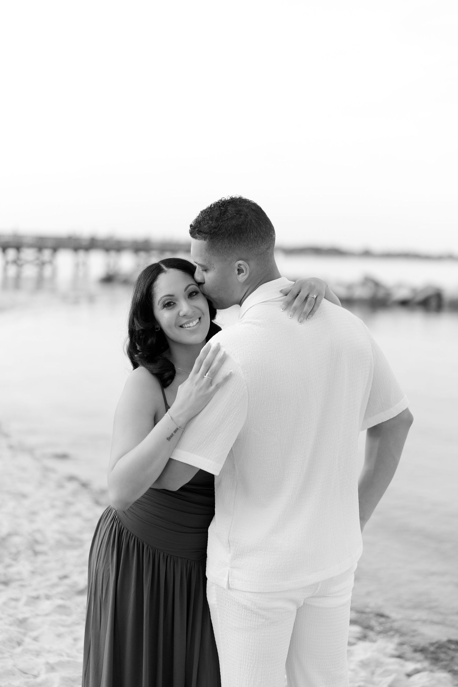Black and white photo of a couple embracing on a beach, woman smiling at camera.