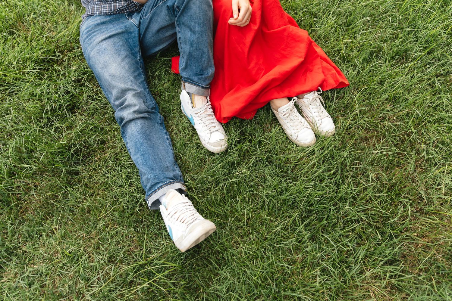 Couple sitting on green grass, wearing jeans and white sneakers, with a red dress visible.