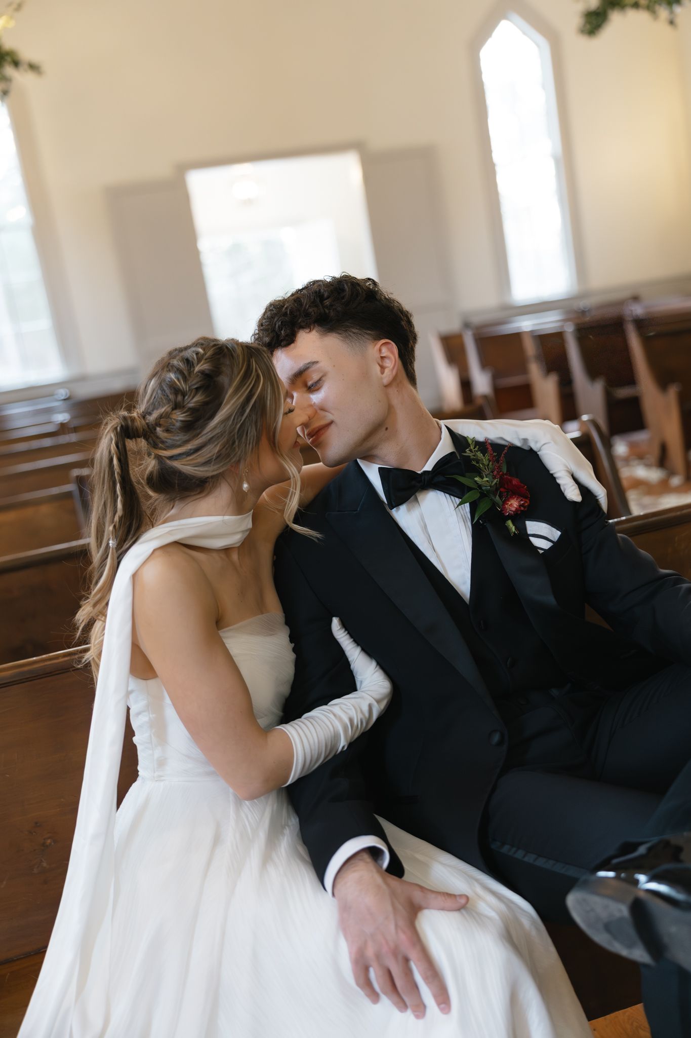 Bride and groom share an intimate moment seated in a church pew, groom in black tuxedo with boutonniere.